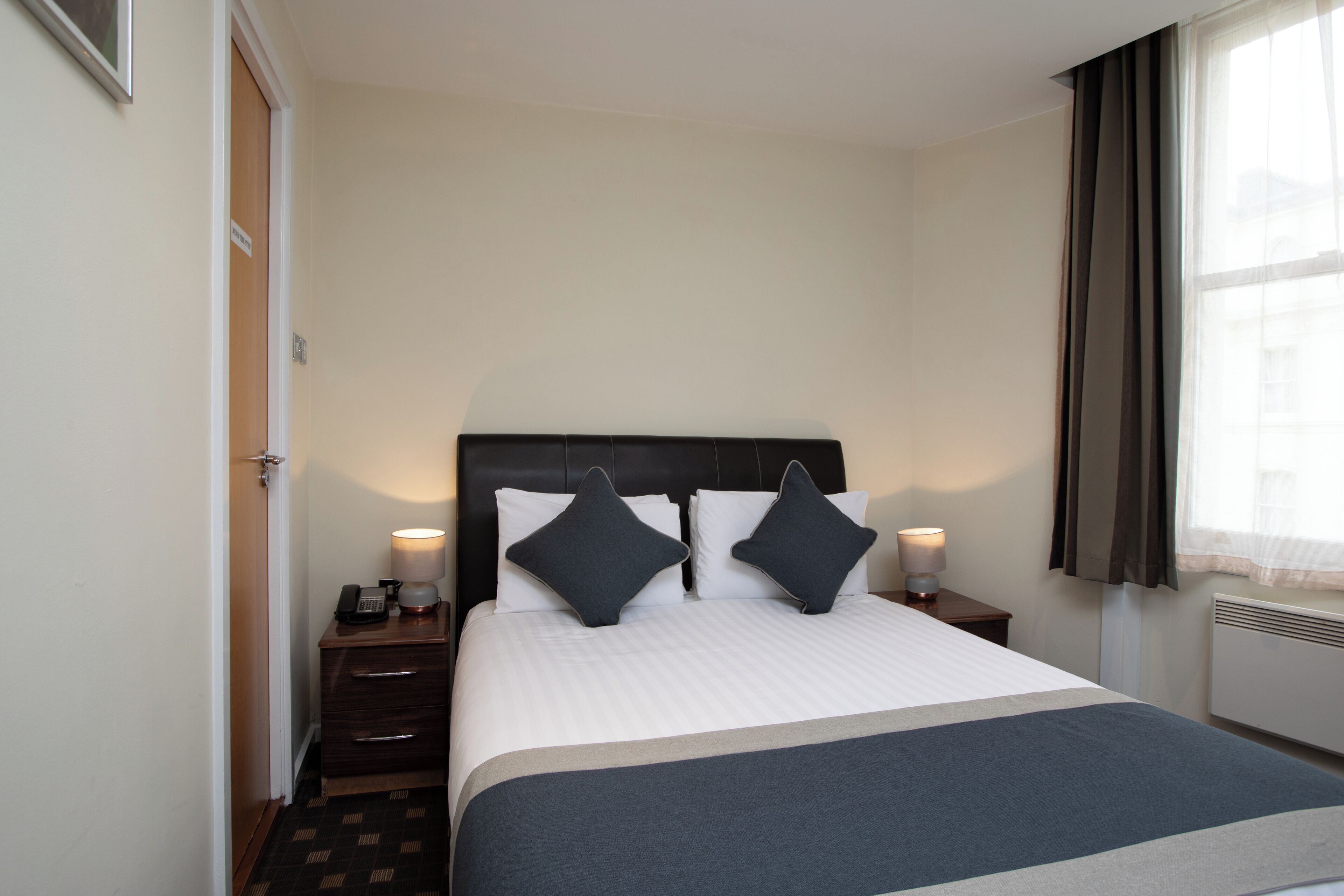 A hotel room with a large bed, two bedside tables, a nightstand, a lamp, and a window with curtains.