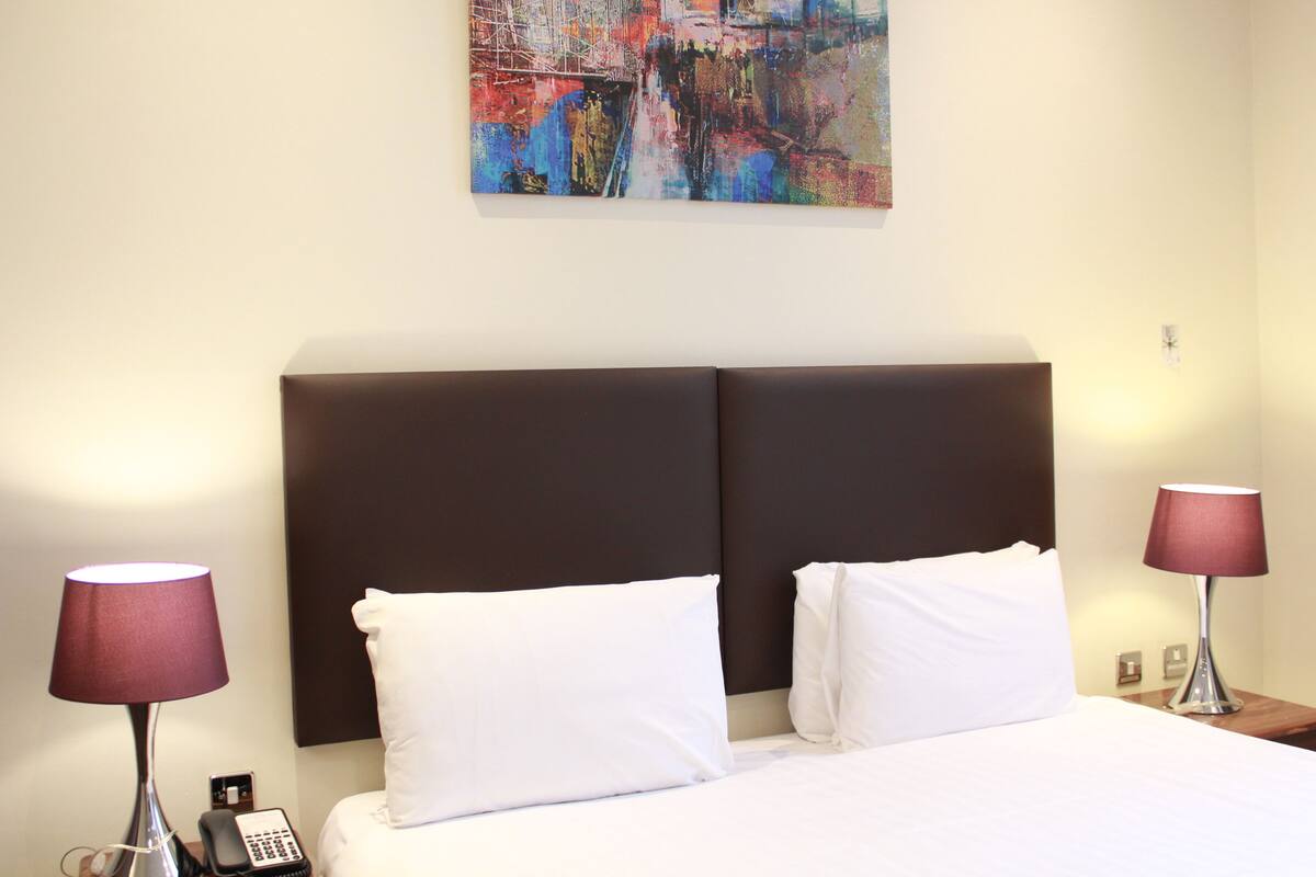Hotellihuone, jossa on suuri sänky, kaksi yöpöytälamppua, puhelin ja värikäs abstrakti maalaus seinällä.