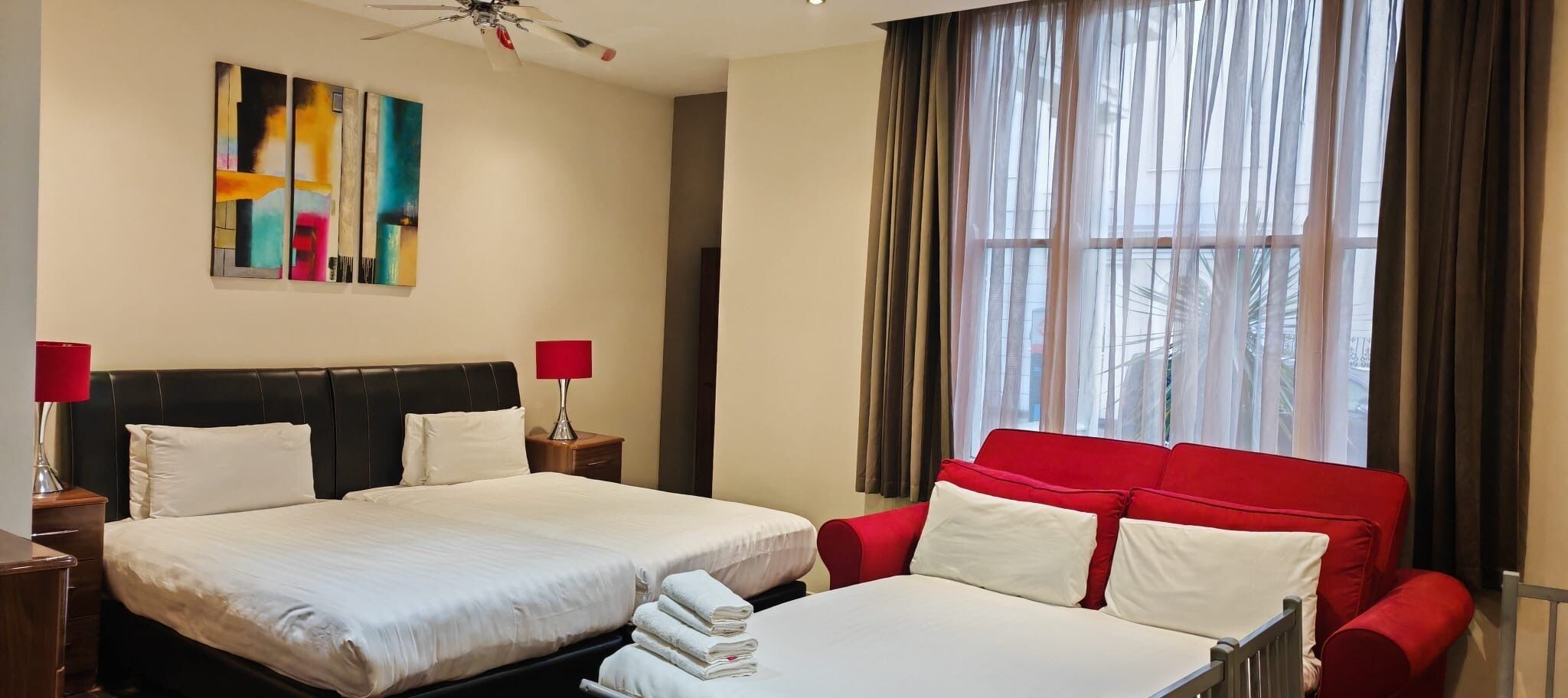 Ein Hotelzimmer mit zwei Betten, einem roten Sofa, einem Deckenventilator und einem großen Fenster mit Vorhängen.