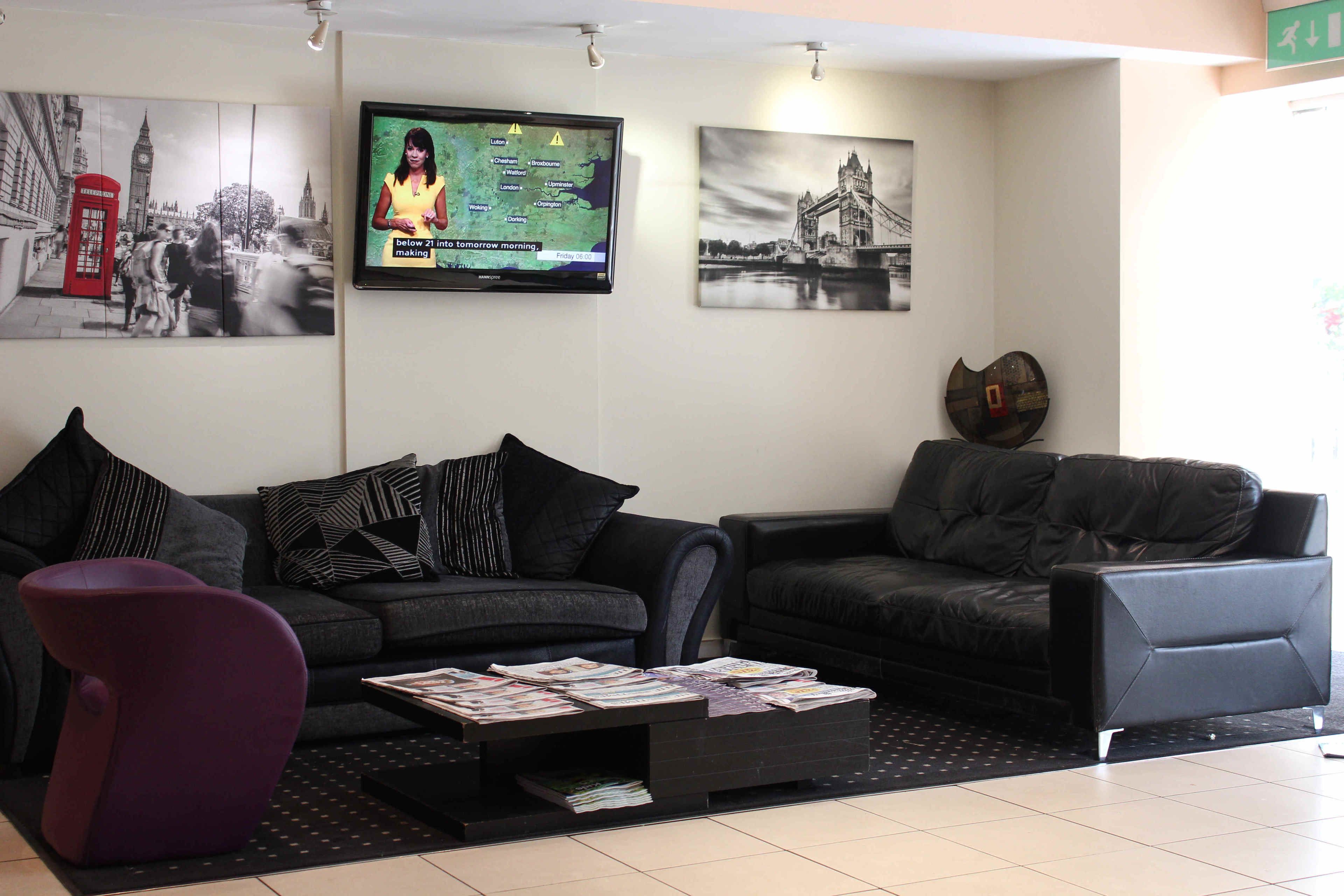 A modern living room with two black sofas, a purple chair, and a coffee table with magazines. There are wall-mounted artworks depicting London landmarks and a television displaying a map.