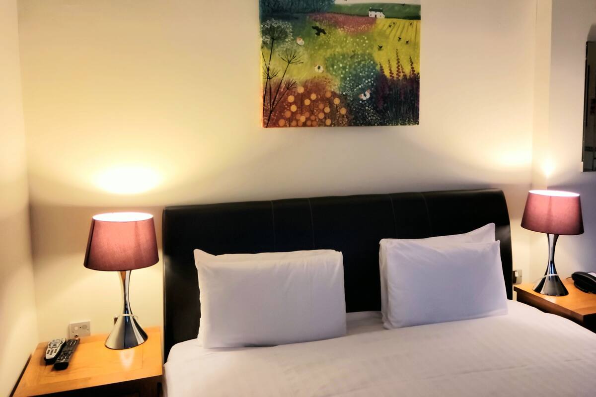 Hotellihuone, jossa on sänky, kaksi yöpöytävalaisinta, taulu seinällä ja kaukosäädin yöpöydällä.