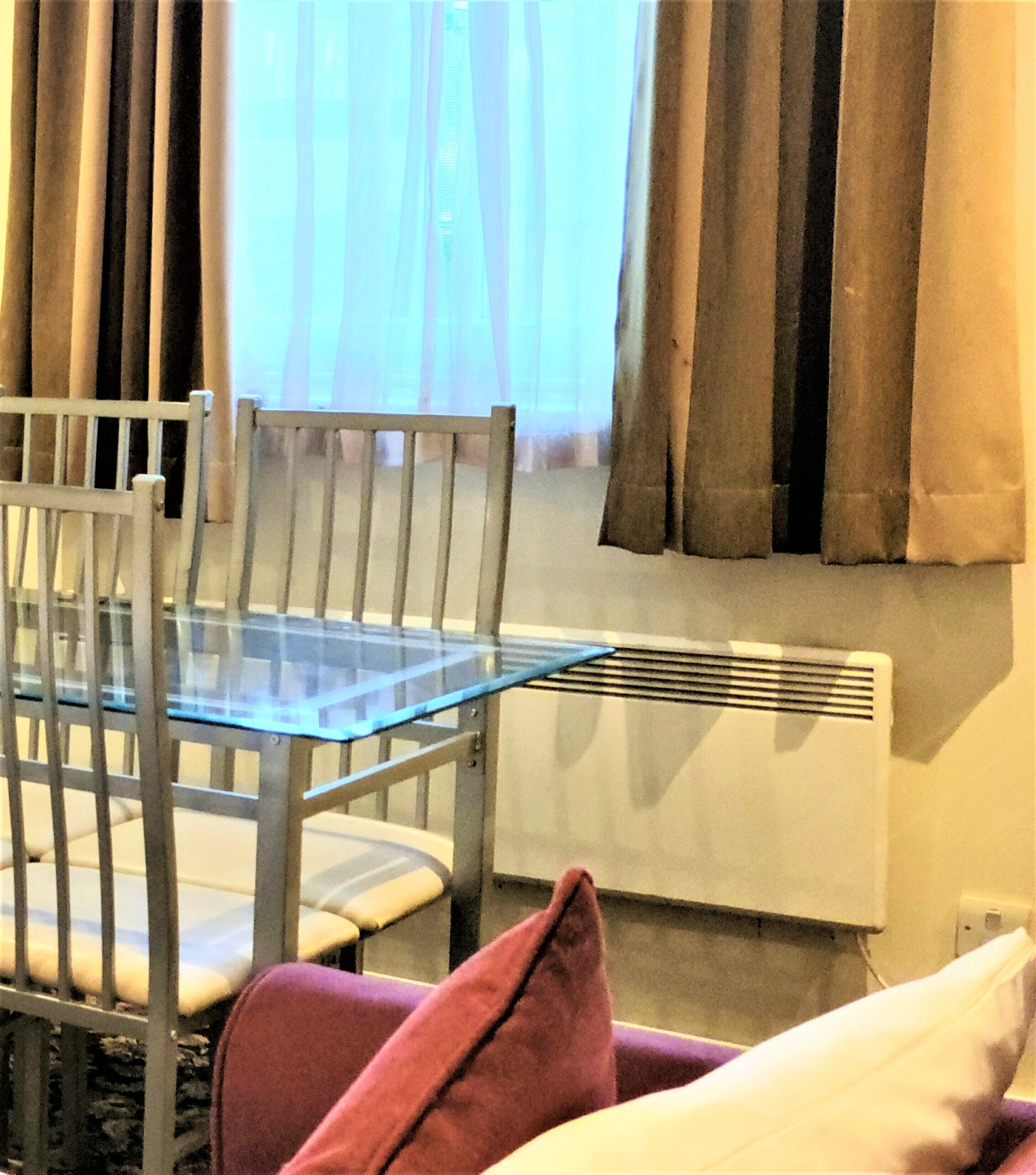 Une pièce avec une table à plateau en verre, une chaise blanche, un coussin rouge, un radiateur blanc et une fenêtre avec des rideaux.