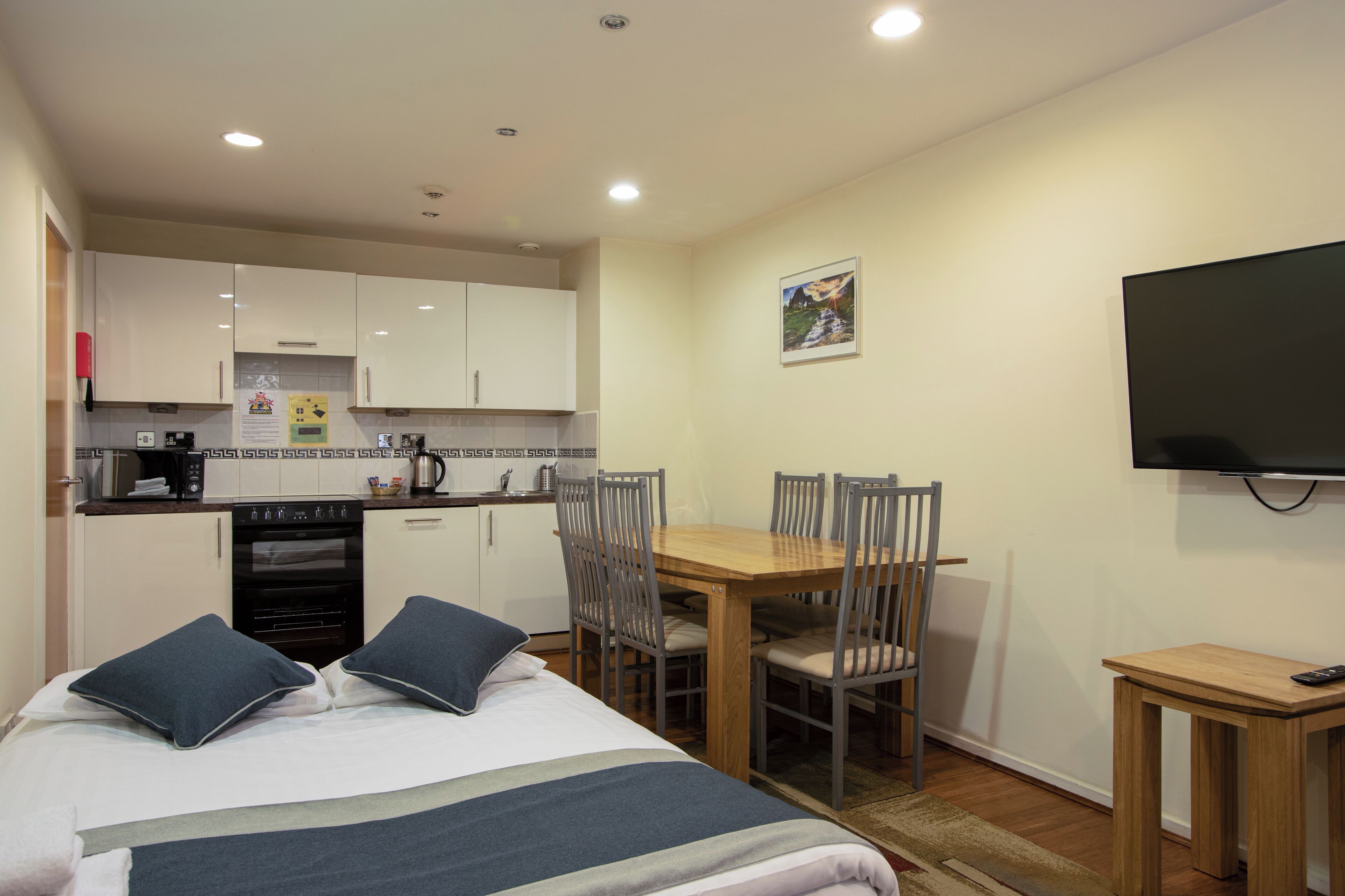 Une chambre d’hôtel compacte comprenant un lit, une table à manger avec des chaises, un petit coin cuisine et un téléviseur fixé au mur.