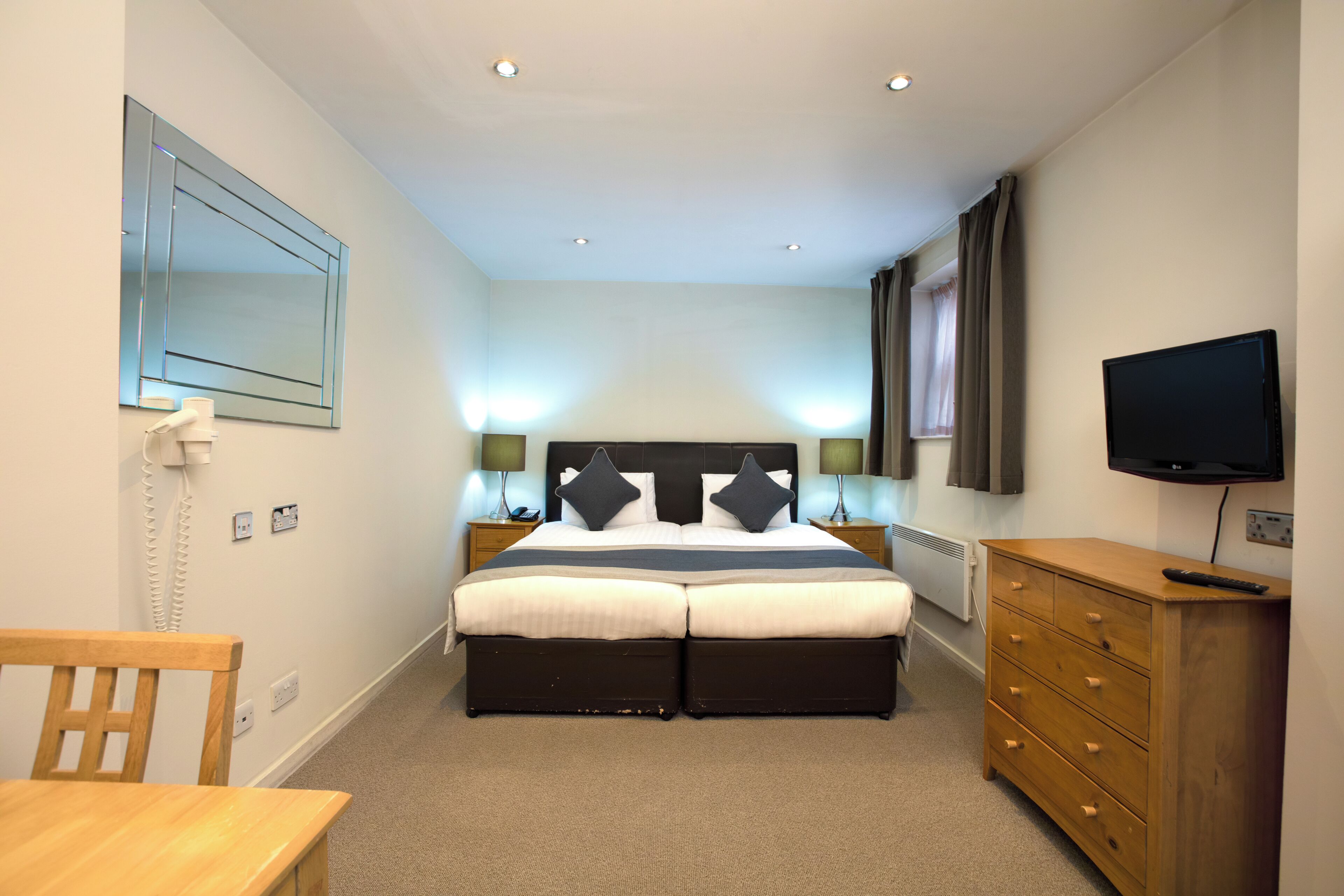 A hotel room with a double bed, bedside lamps, a TV, a wooden dresser, and a desk with a chair.