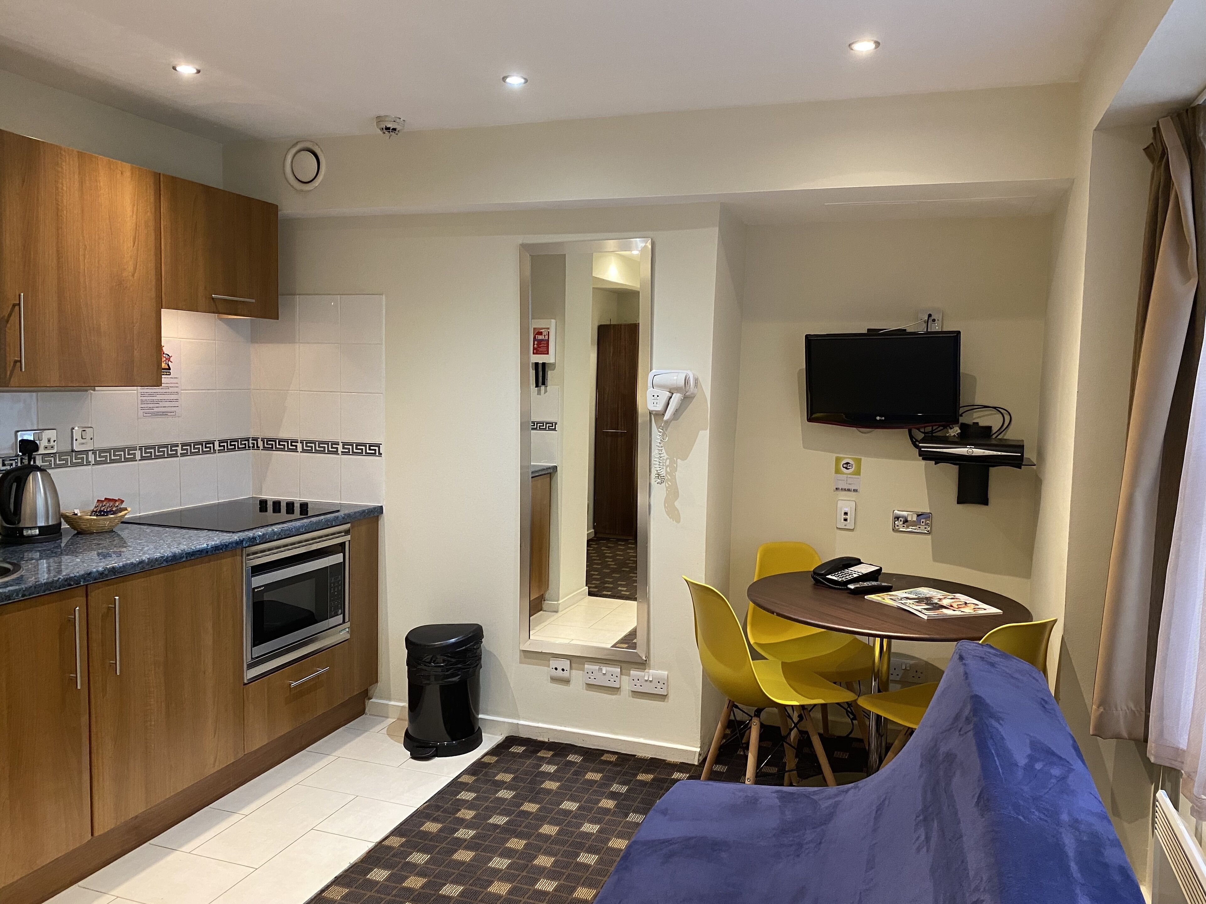 A compact hotel room with a kitchenette, a dining area with yellow chairs, a blue sofa, and a flat-screen TV mounted on the wall.