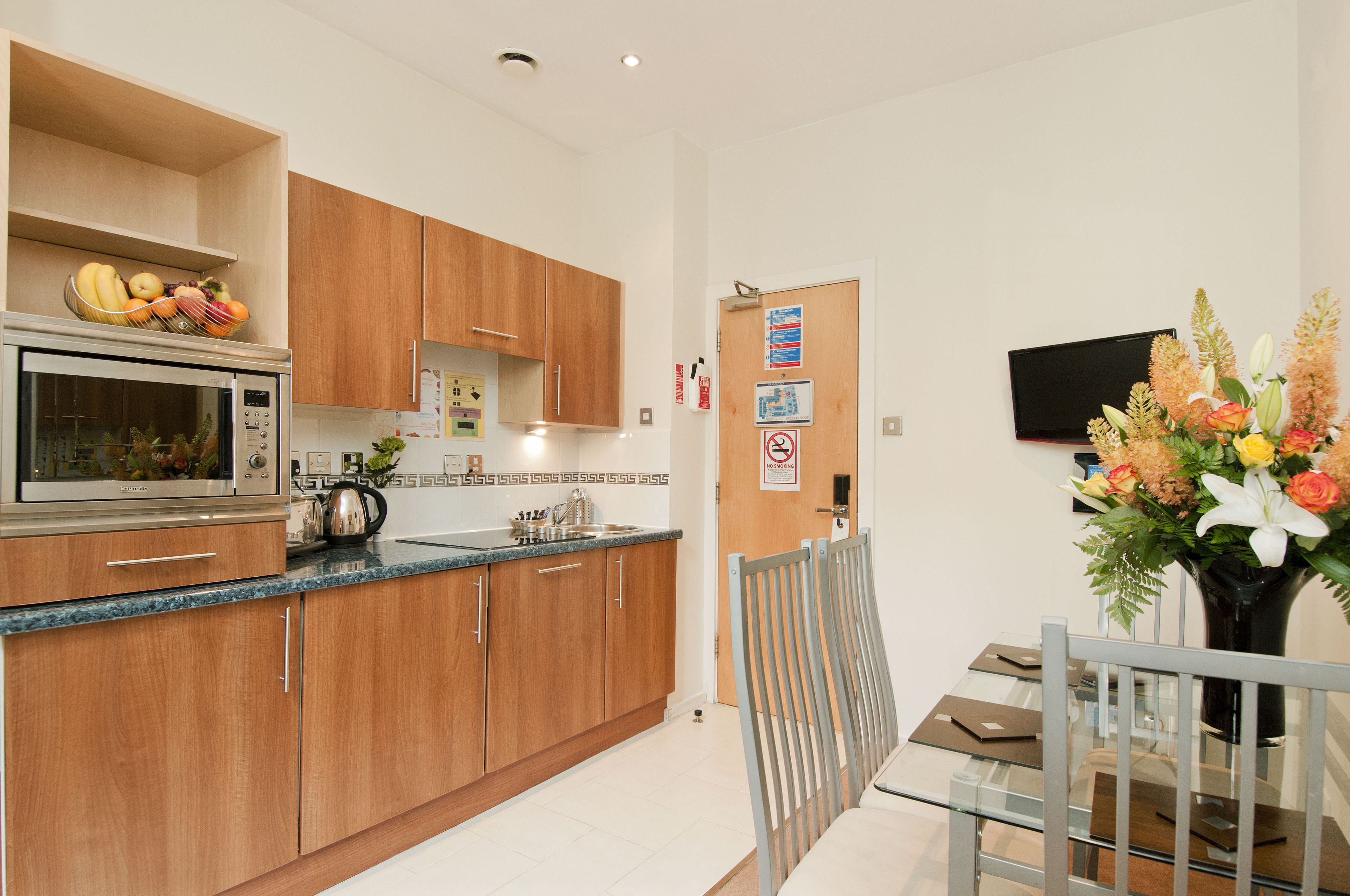 A modern kitchen with wooden cabinets, a stainless steel microwave, and a dining area with a glass table and metal chairs.