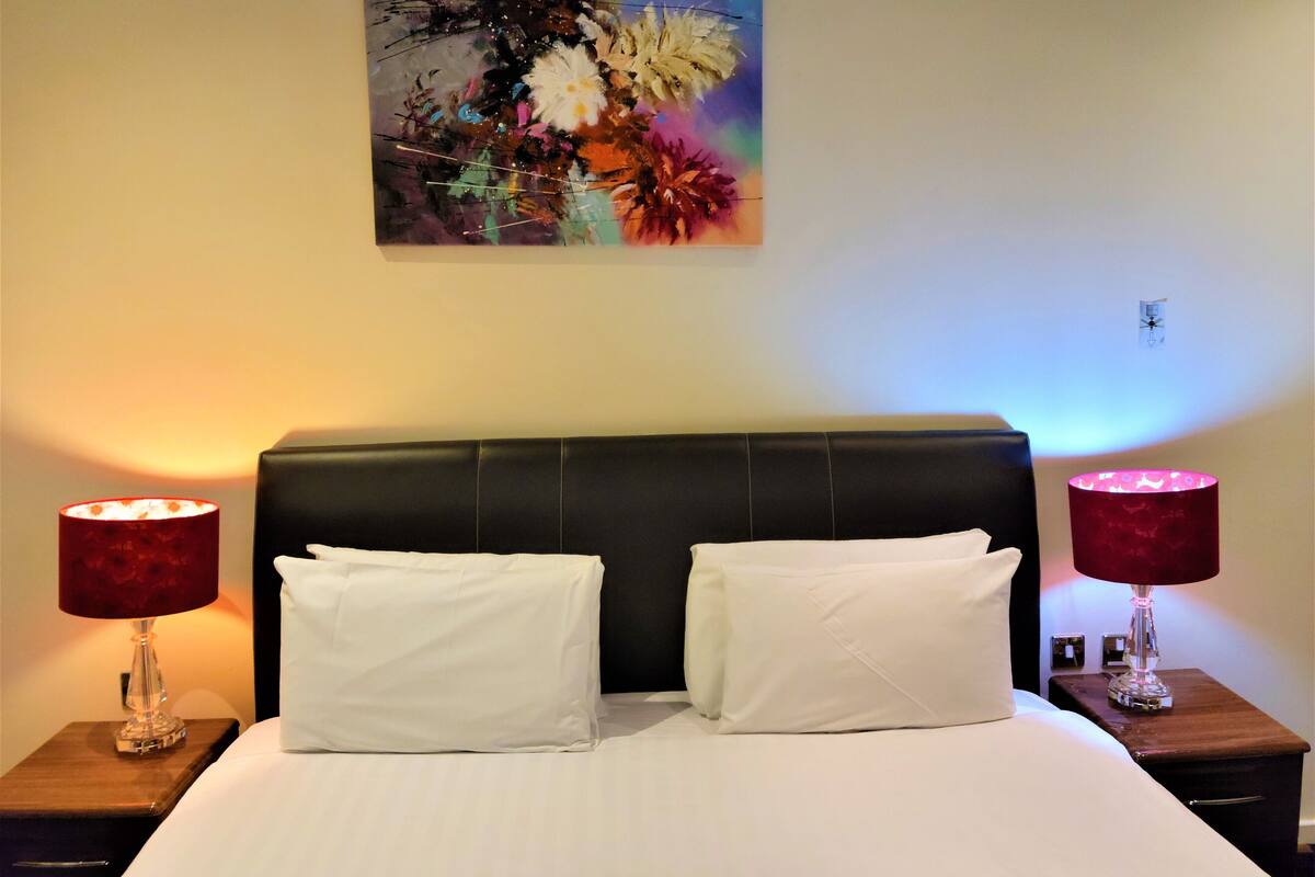 Hotellihuone, jossa on suuri sänky, kaksi yöpöytää ja seinälle ripustettu taulu.