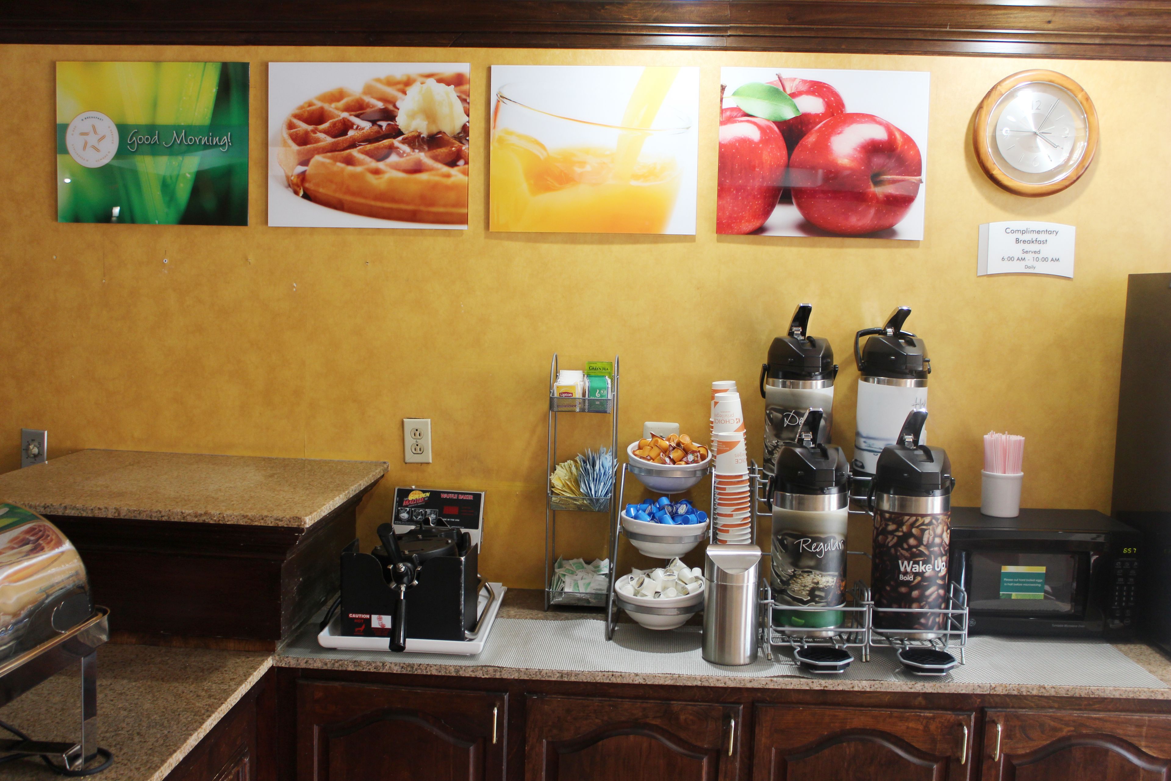 Una barra de desayuno con gofres, jugo de naranja y manzanas. Hay cafeteras, un microondas y un reloj en la pared.
