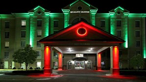 Entrada de hotel con fachada iluminada en rojo y verde, acceso despejado y un cartel que dice "QUALITY SUITES".