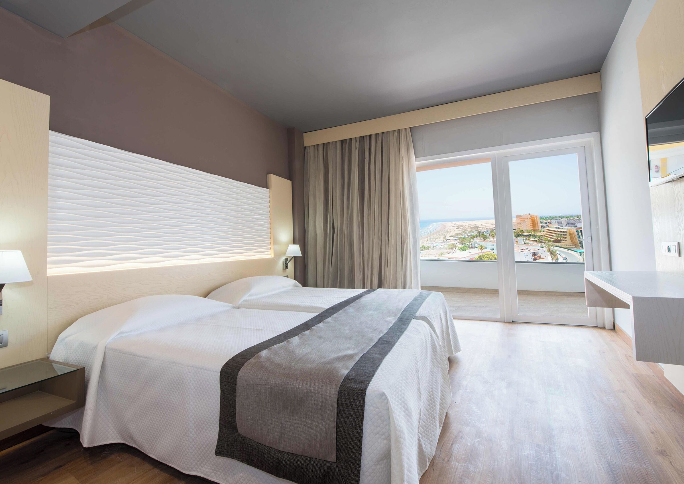 A hotel room with a bed, bedside table, lamp, and a view of the city through a large window.
