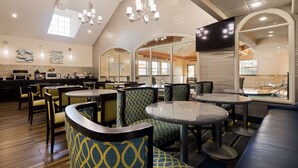 A modern restaurant with round tables and patterned chairs, a kitchen area, and a pool view. - Best Western Plus Tulsa Inn & Suites (Tulsa)