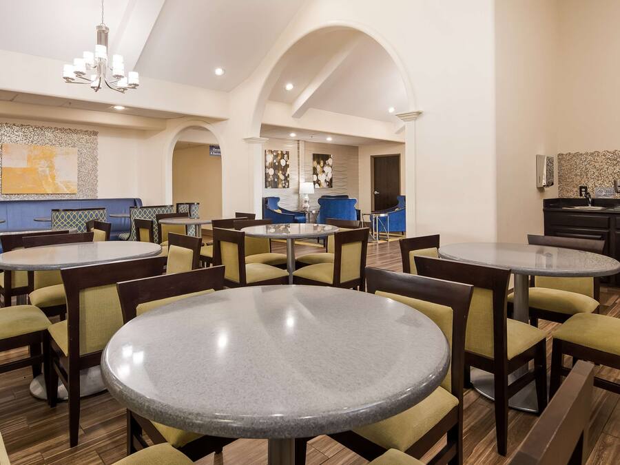 A hotel dining area with round tables and chairs, a chandelier, and a kitchen area visible in the background.