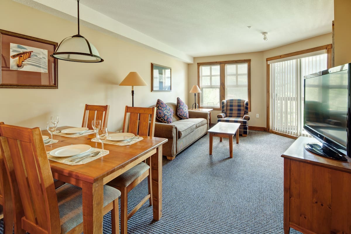 Una sala de estar con una mesa de comedor para cuatro personas, un sofá, un televisor y ventanas grandes.