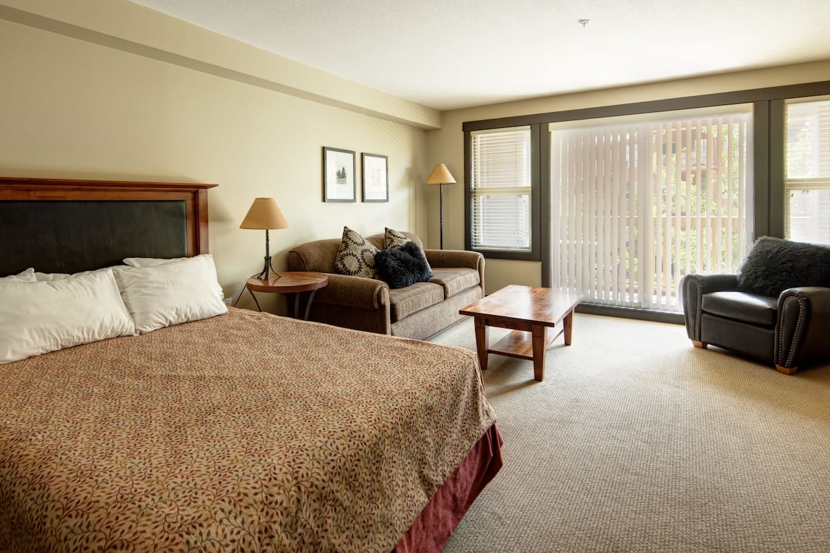 Habitación de hotel con una cama grande, un sofá, una silla, una mesita y una ventana con persianas.