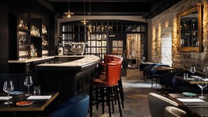 Un espace bar avec un comptoir, des tabourets et un présentoir à vin. On y trouve des tables avec des chaises et des verres à vin. La pièce est dotée d’un mur en pierre et d’un grand miroir.