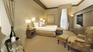 Une chambre d’hôtel comprenant un lit, deux lampes de chevet, une table de chevet, une commode, une chaise, une petite table avec une bouteille et des verres, et une fenêtre avec des rideaux.