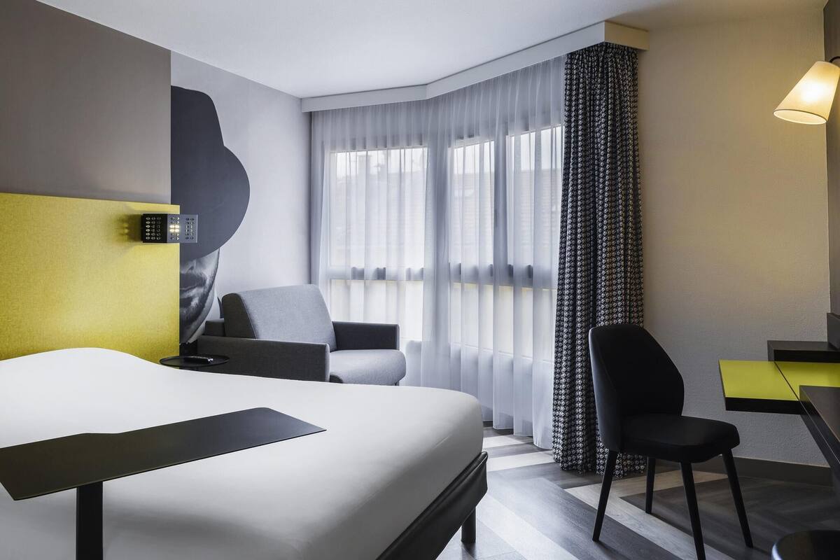 Una camera d'albergo con un letto, due poltrone, una scrivania, una lampada e un'ampia finestra con tende leggere.