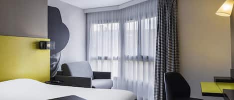 Habitación de hotel con cama, dos sillas, un escritorio, una lámpara y un gran ventanal con cortinas traslúcidas.
