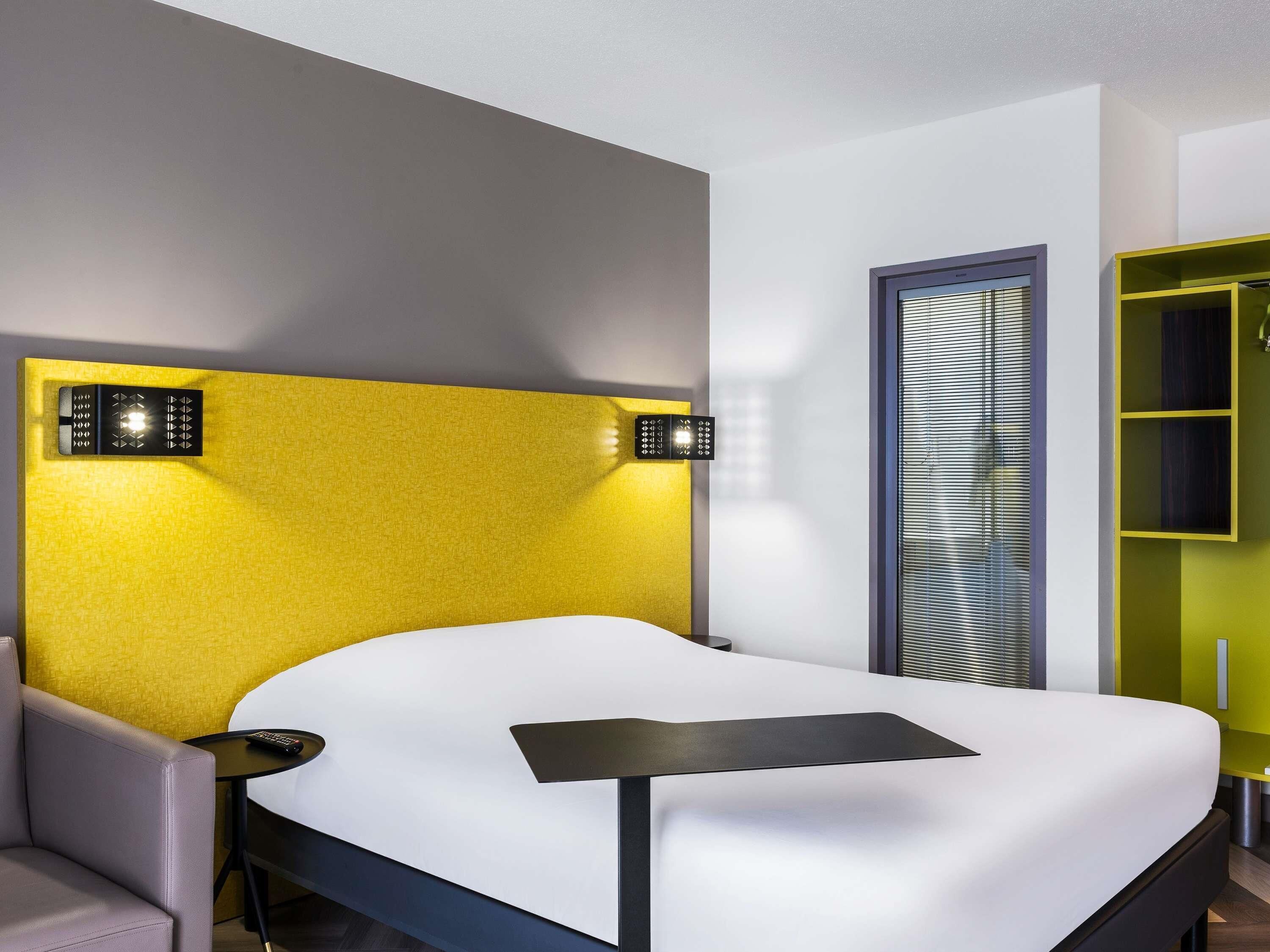A modern hotel room with a bed, a small table, a chair, and a built-in wardrobe.
