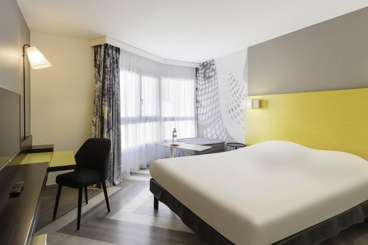 A hotel room with a bed, a desk, a chair, a small table, and a window with curtains.