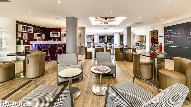 A modern hotel lobby with a central reception desk, multiple seating areas with tables and chairs, and a ceiling fan. - ibis Styles Nice Vieux Port (Nice)