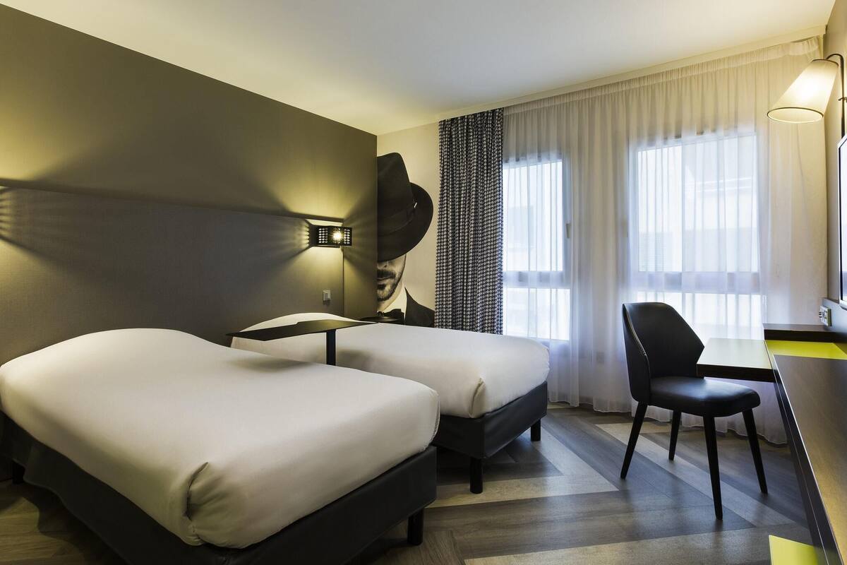 Camera d'albergo con due letti, una scrivania, una sedia, una lampada a parete e un'ampia finestra con tende.