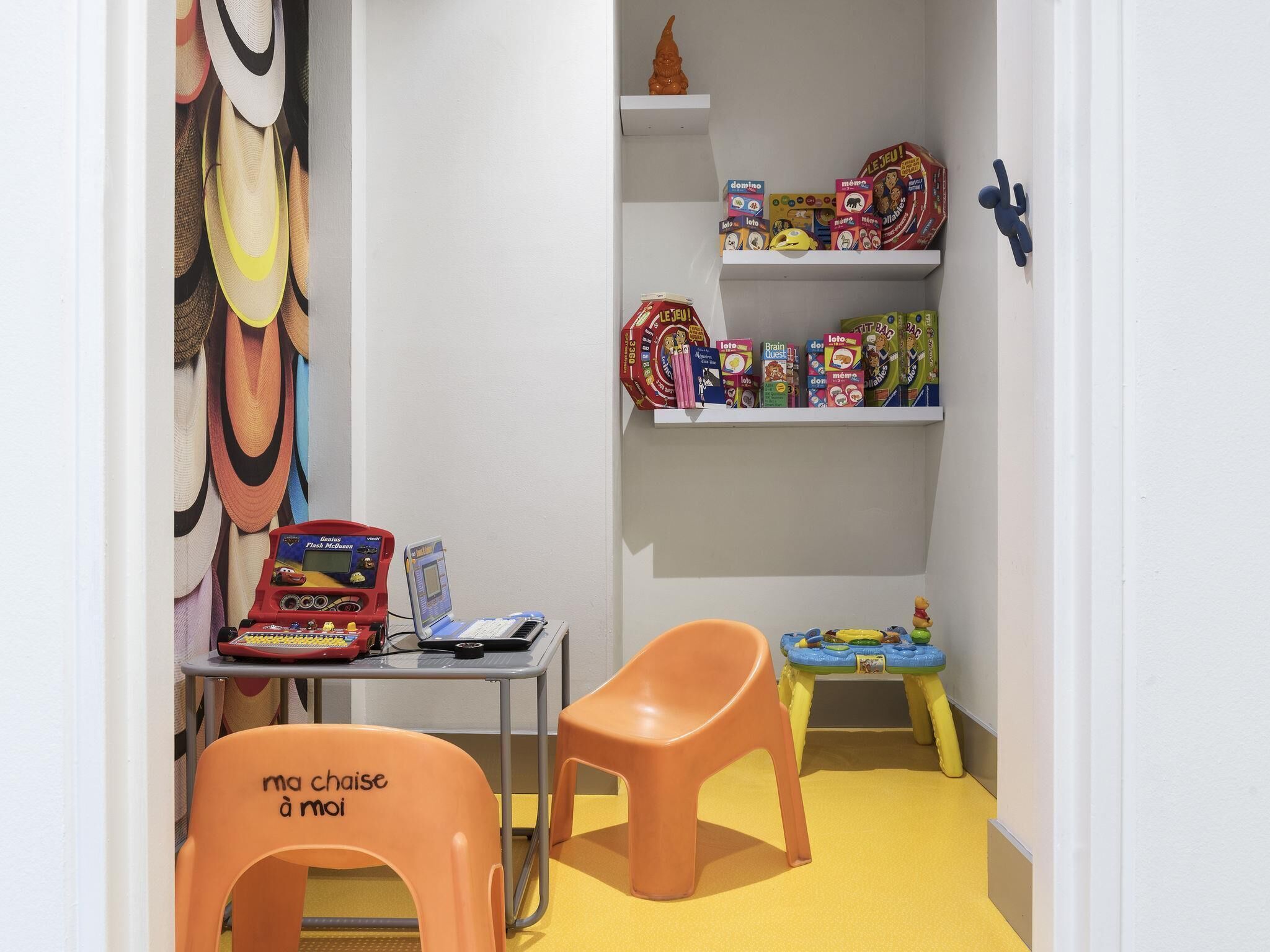 Espaço infantil com brinquedos, uma mesa pequena e cadeiras laranjas.