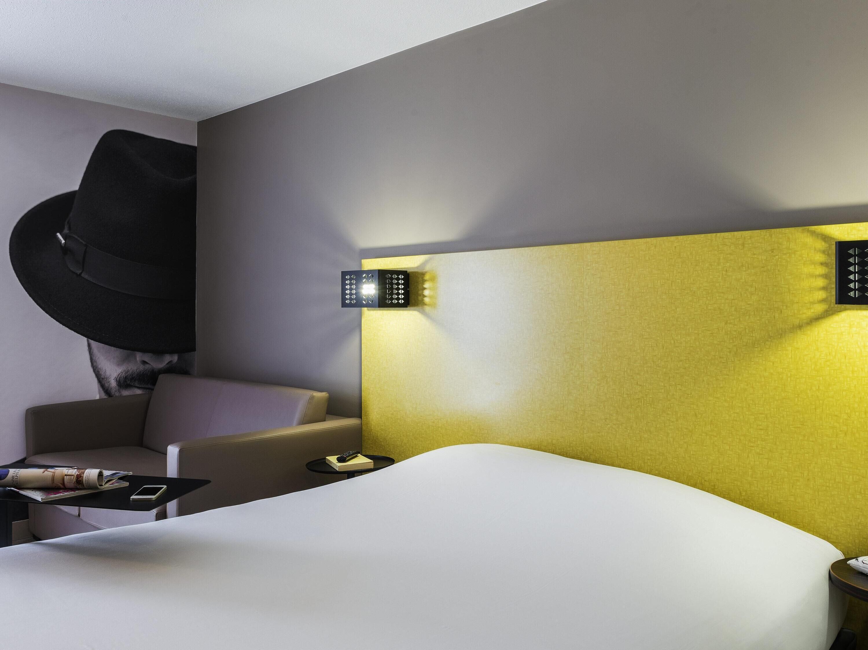 A modern hotel room with a curved bed, a headboard with integrated lighting, a small round table with a remote control, and a wall-mounted hat rack.