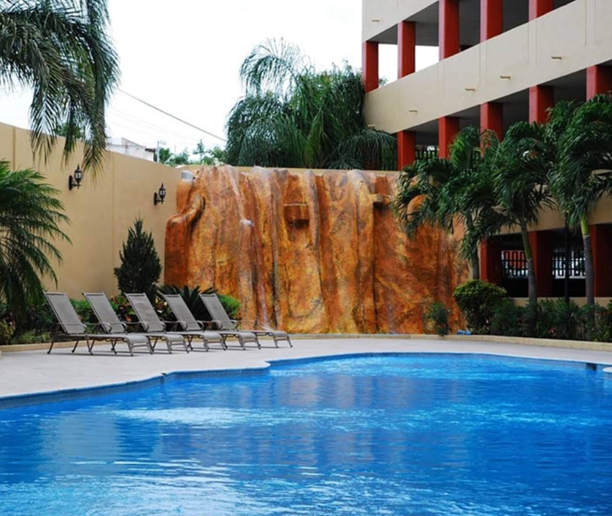 A swimming pool with a waterfall feature, surrounded by lounge chairs and palm trees.