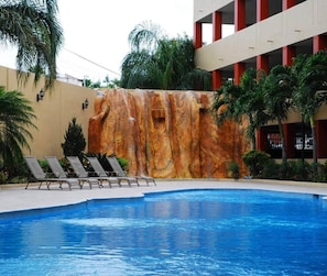 A swimming pool with a waterfall feature, surrounded by lounge chairs and palm trees. - Royal Garden Reynosa (Reynosa)