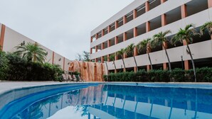 Una piscina con reflejo cristalino, rodeada de palmeras y un edificio de varios pisos con balcones.