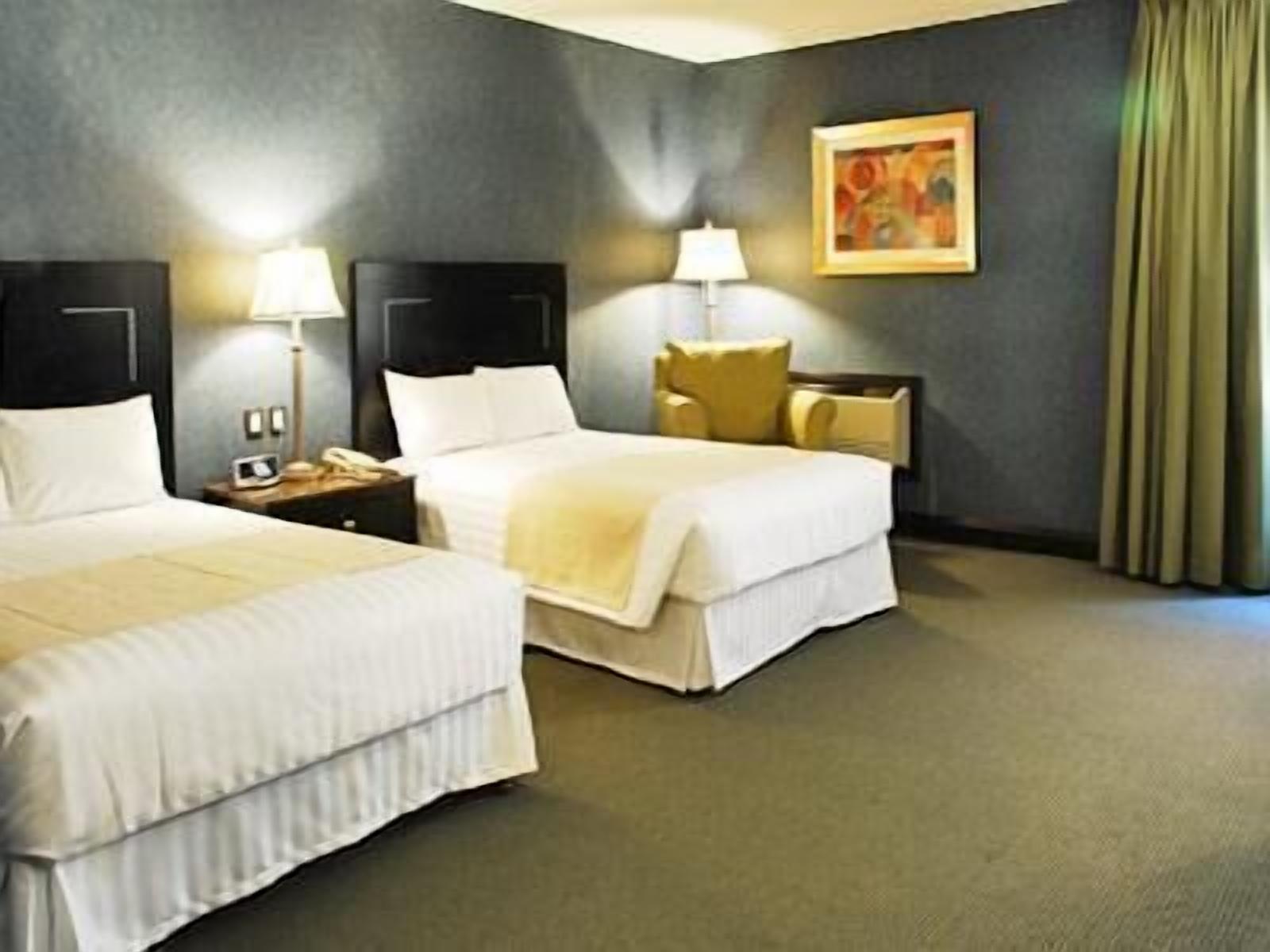A hotel room with two beds, a nightstand, a lamp, and a painting on the wall.