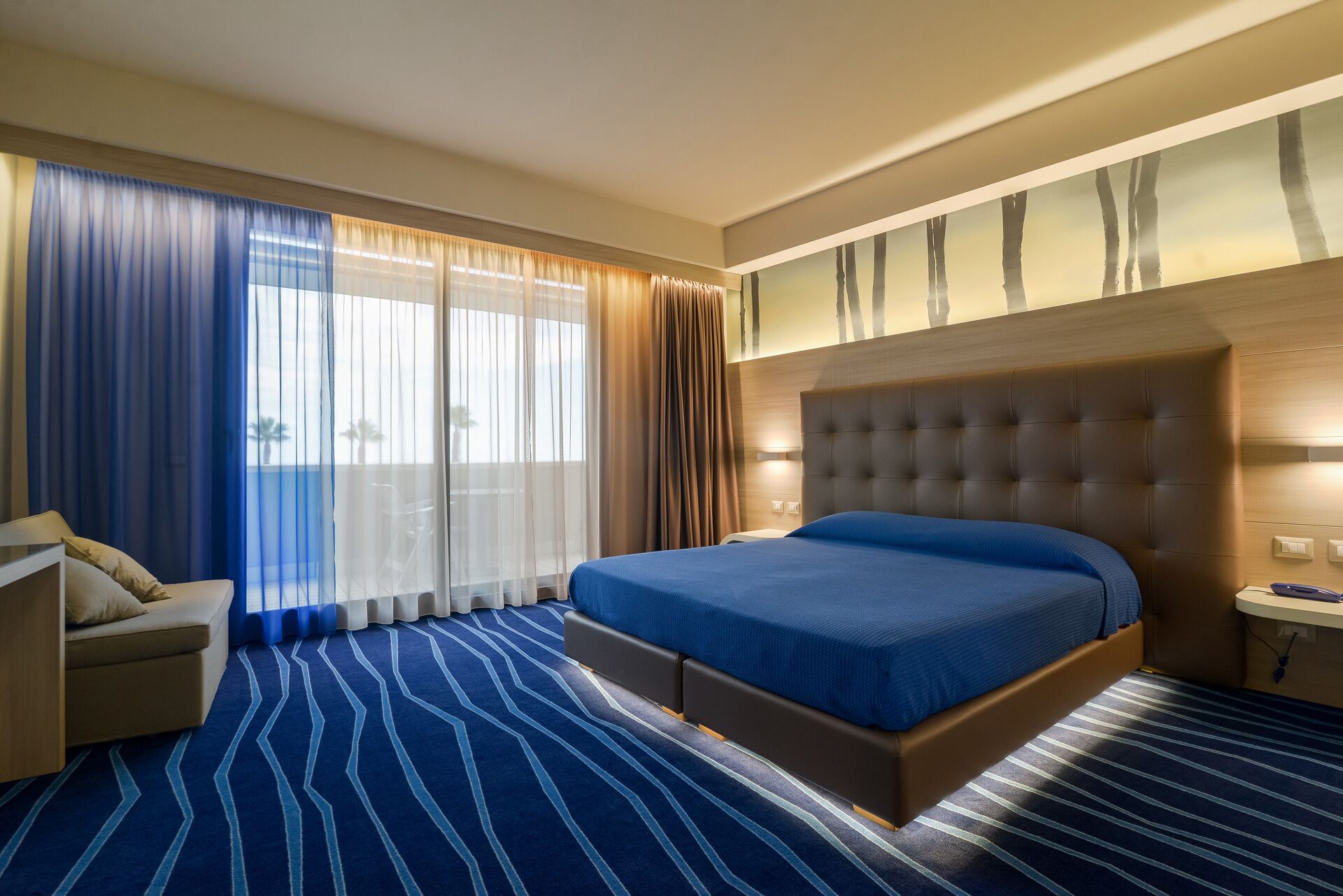 Ein Hotelzimmer mit einem großen Bett, einem Sofa, einem Schreibtisch und Blick auf Palmen durch die Fenster.