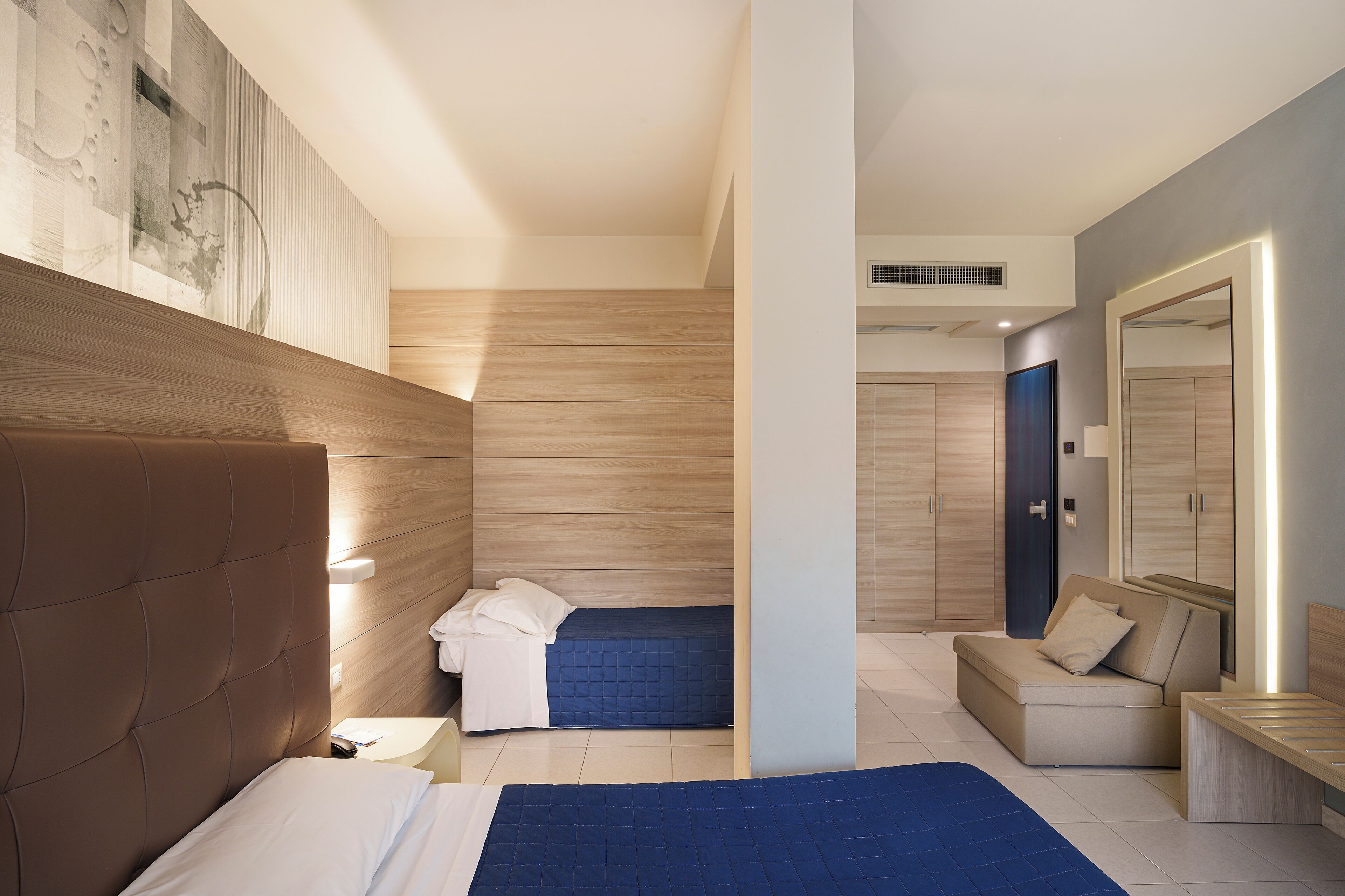 A modern hotel room with a large bed, a smaller bed, a sofa, and a desk.