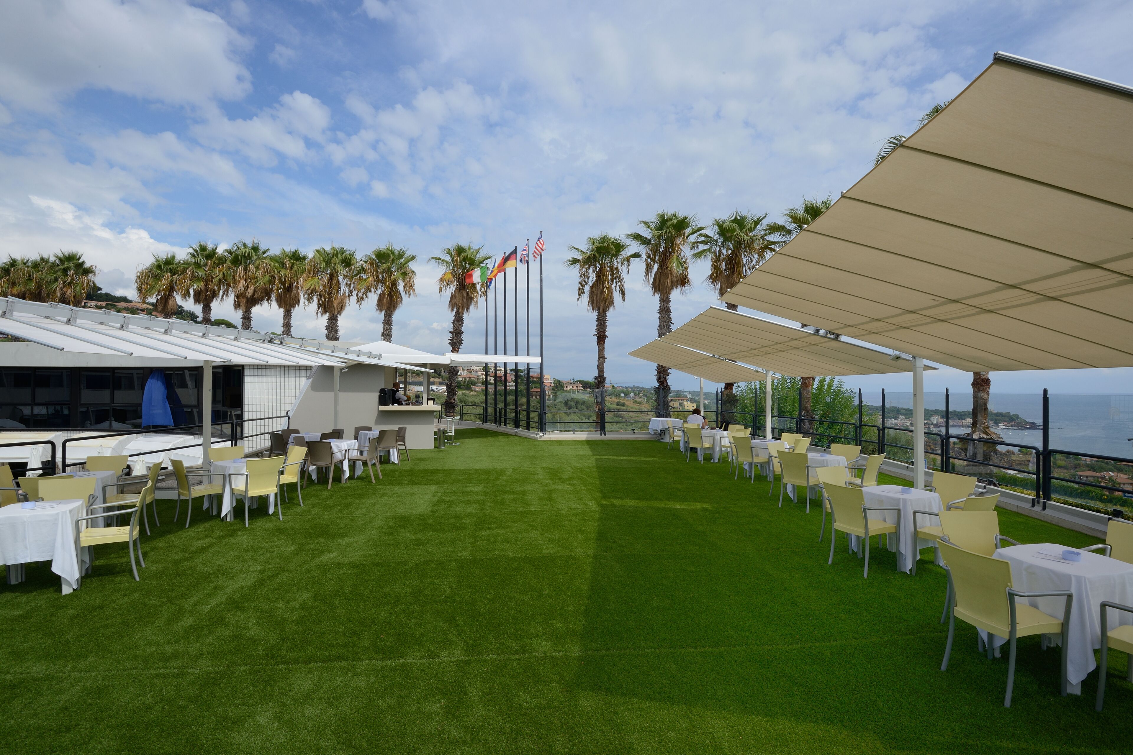 Un área de comedor al aire libre con mesas y sillas, palmeras y banderas.