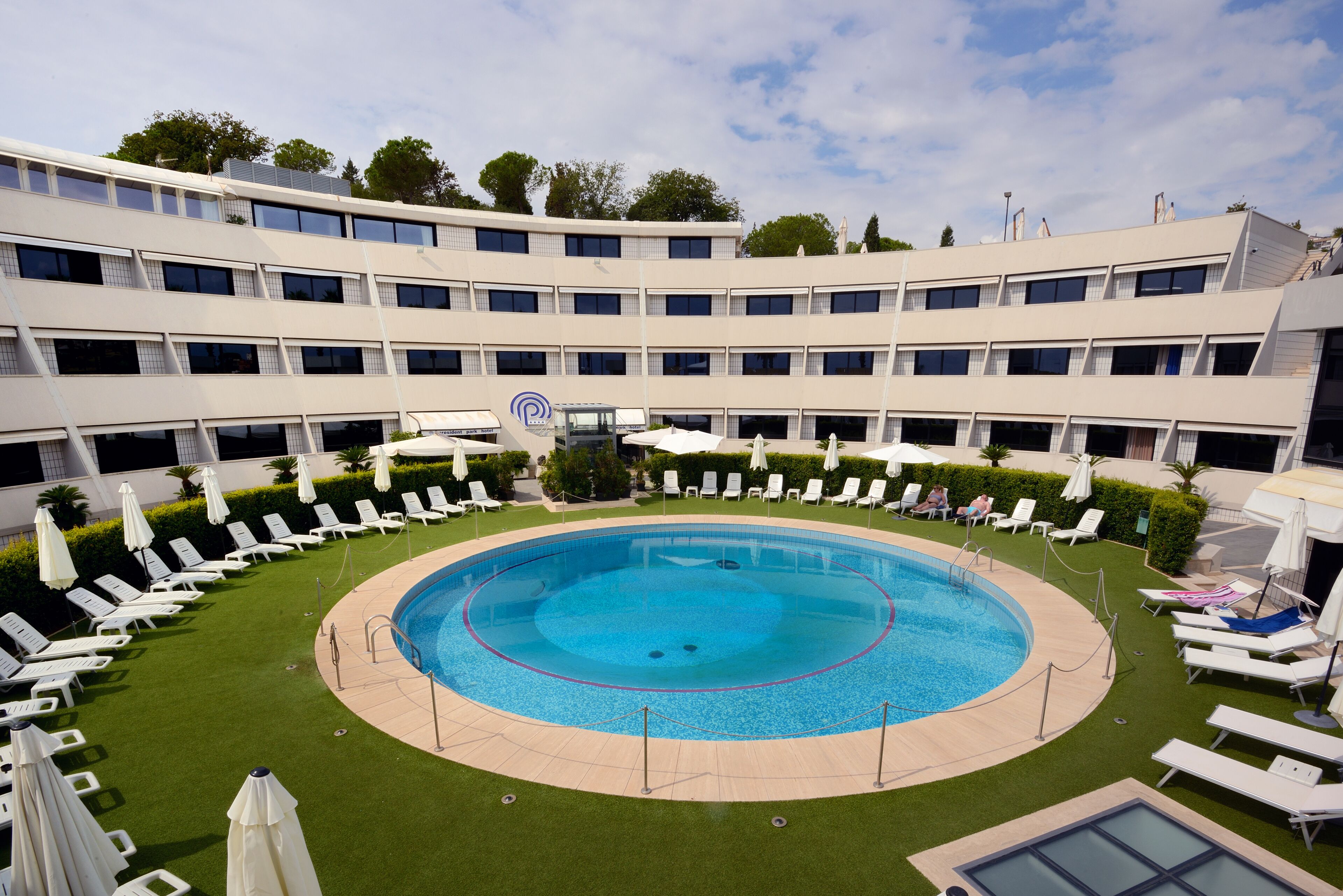 Een hotel met een rond zwembad omringd door ligbedden en parasols, een fontein in het midden en een meerlaags gebouw met balkons.