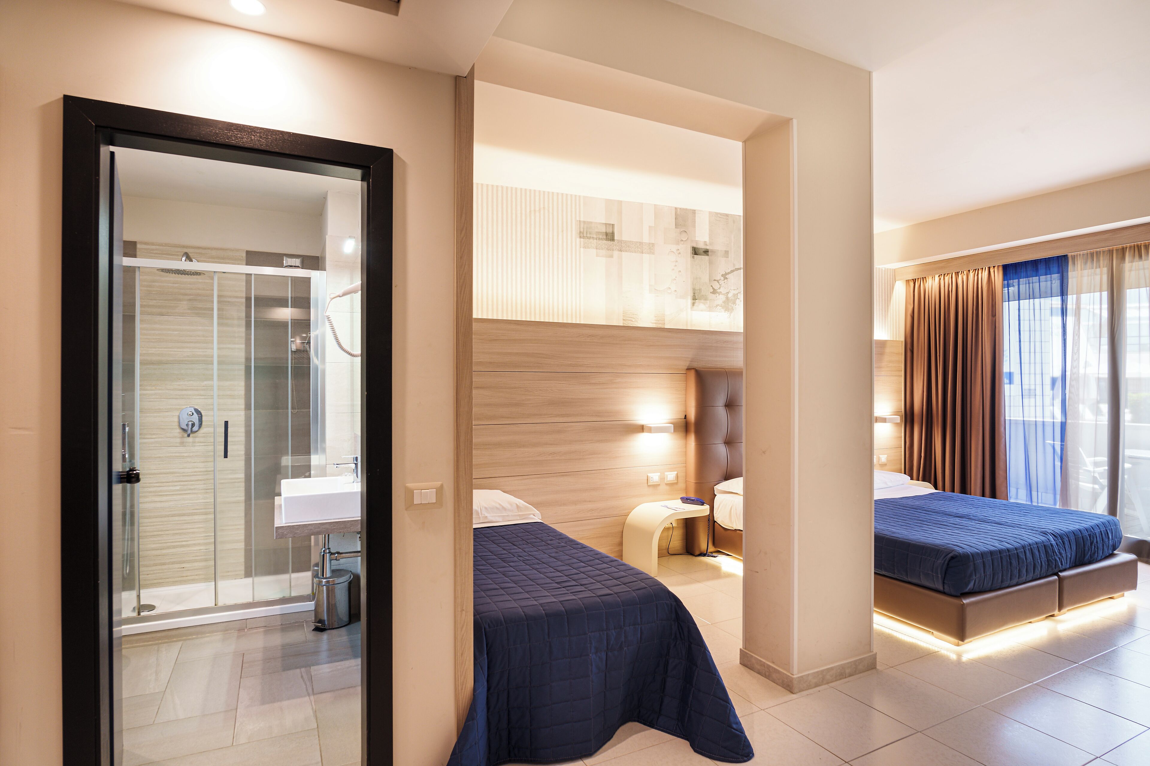 Quarto de hotel moderno com box de vidro, banheiro separado, cama com cabeceira e mesa de cabeceira.