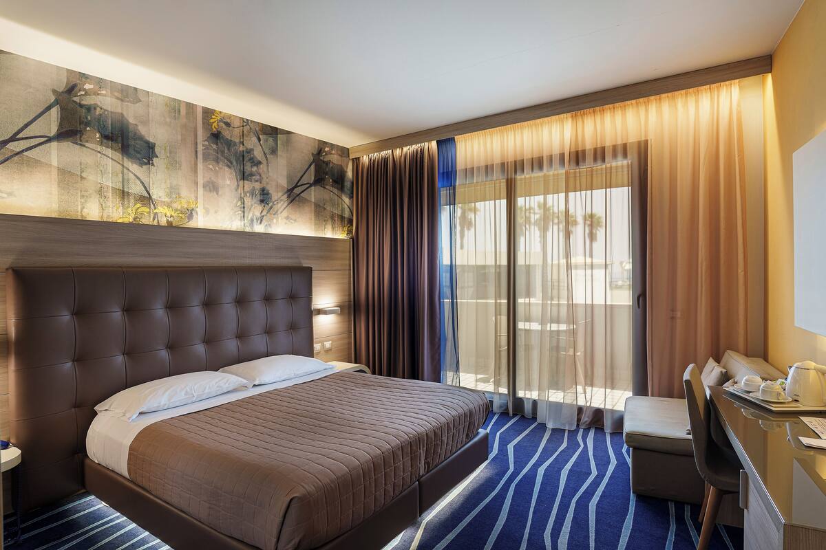 Een hotelkamer met een groot bed, een bureau en uitzicht op palmbomen door dunne gordijnen.