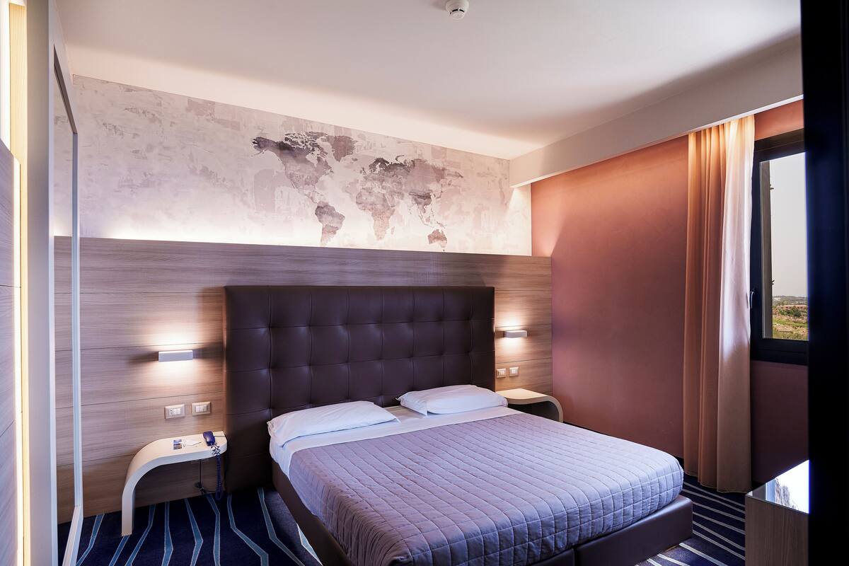 Een moderne hotelkamer met een groot bed, een wereldkaart aan de muur en uitzicht op het omliggende landschap door het raam.