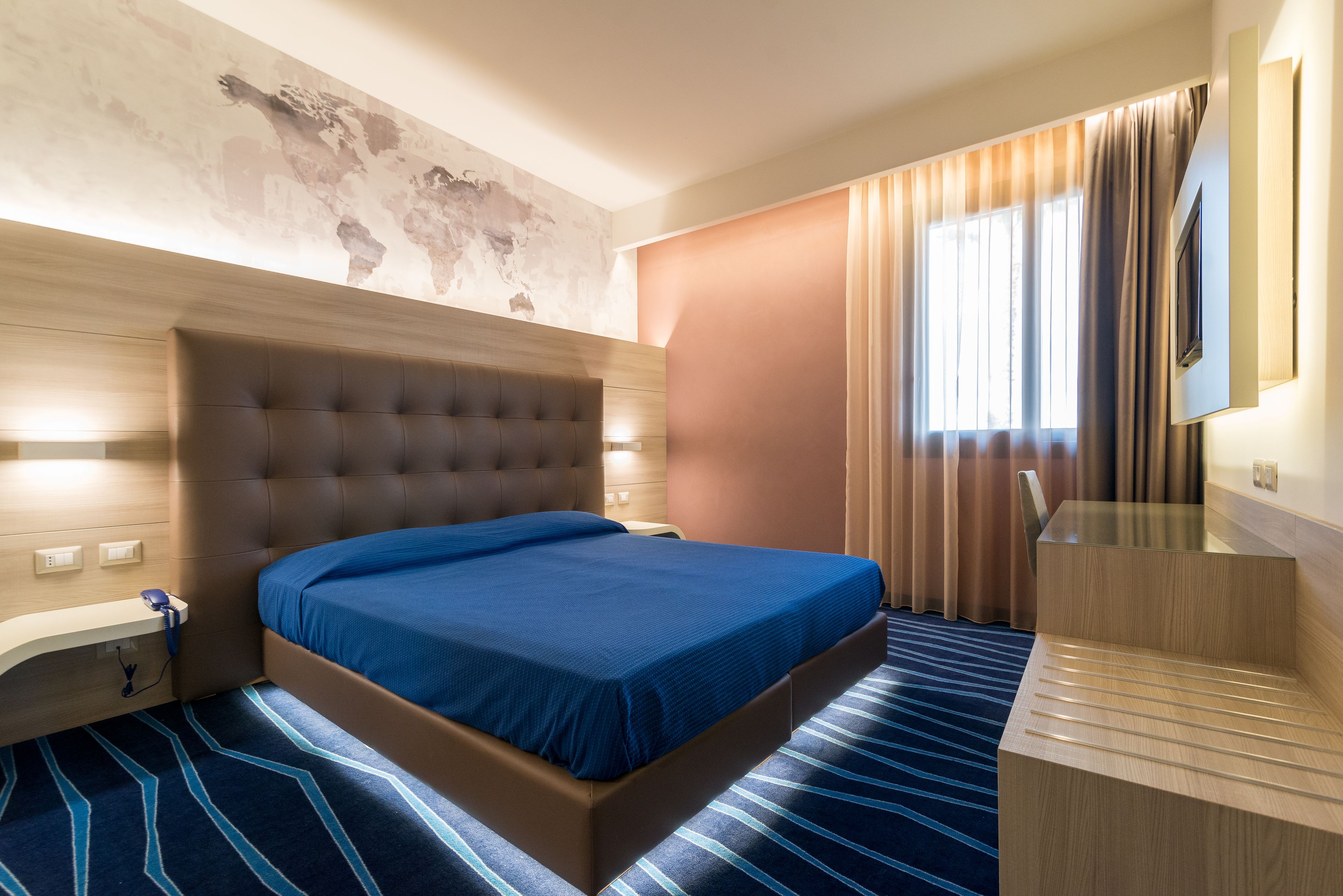 A hotel room with a large bed, a wooden headboard, a desk, and a world map on the wall.