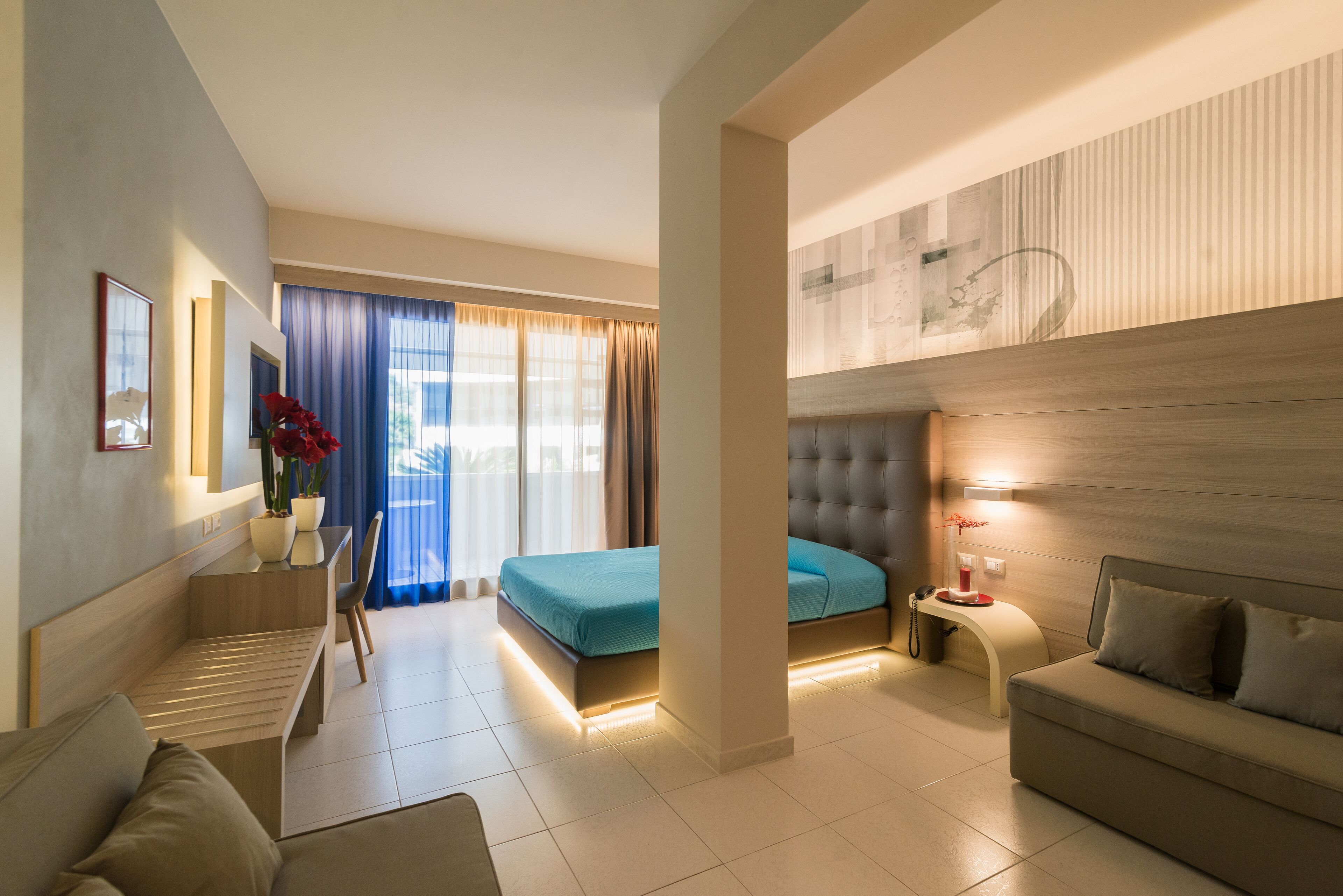 A modern hotel room with a bed, a sofa, a desk, and a small table with a lamp.
