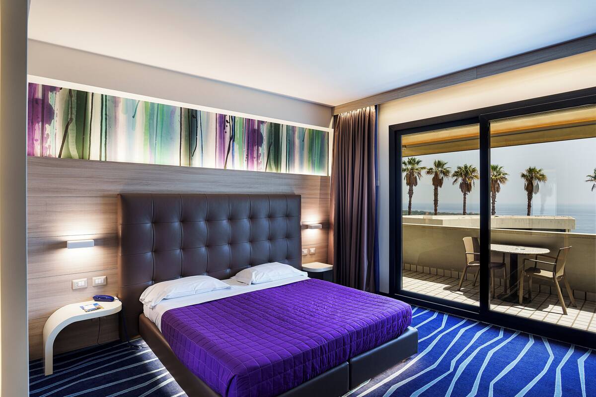 Een moderne hotelkamer met een groot bed, een nachtkastje, een raam met uitzicht op palmbomen en een balkon met buitenmeubilair.