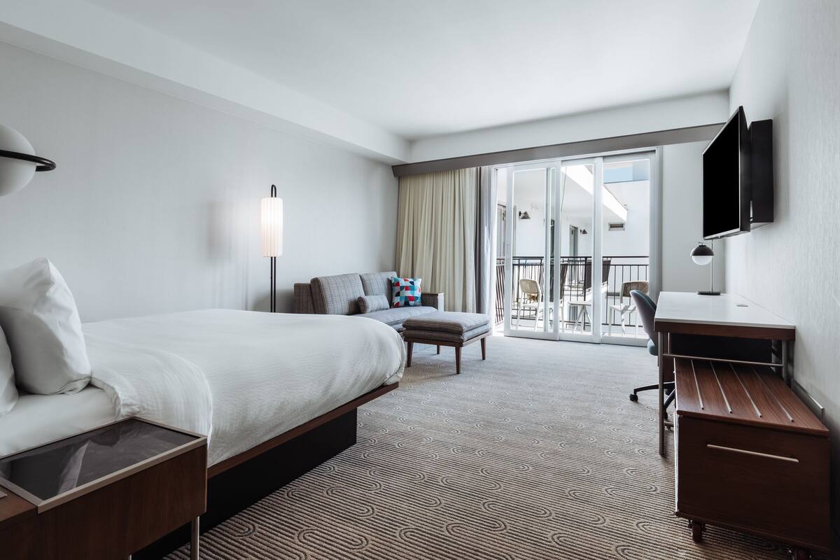 Quarto de hotel com uma cama grande, uma escrivaninha, uma cadeira, televisão e varanda com vista.