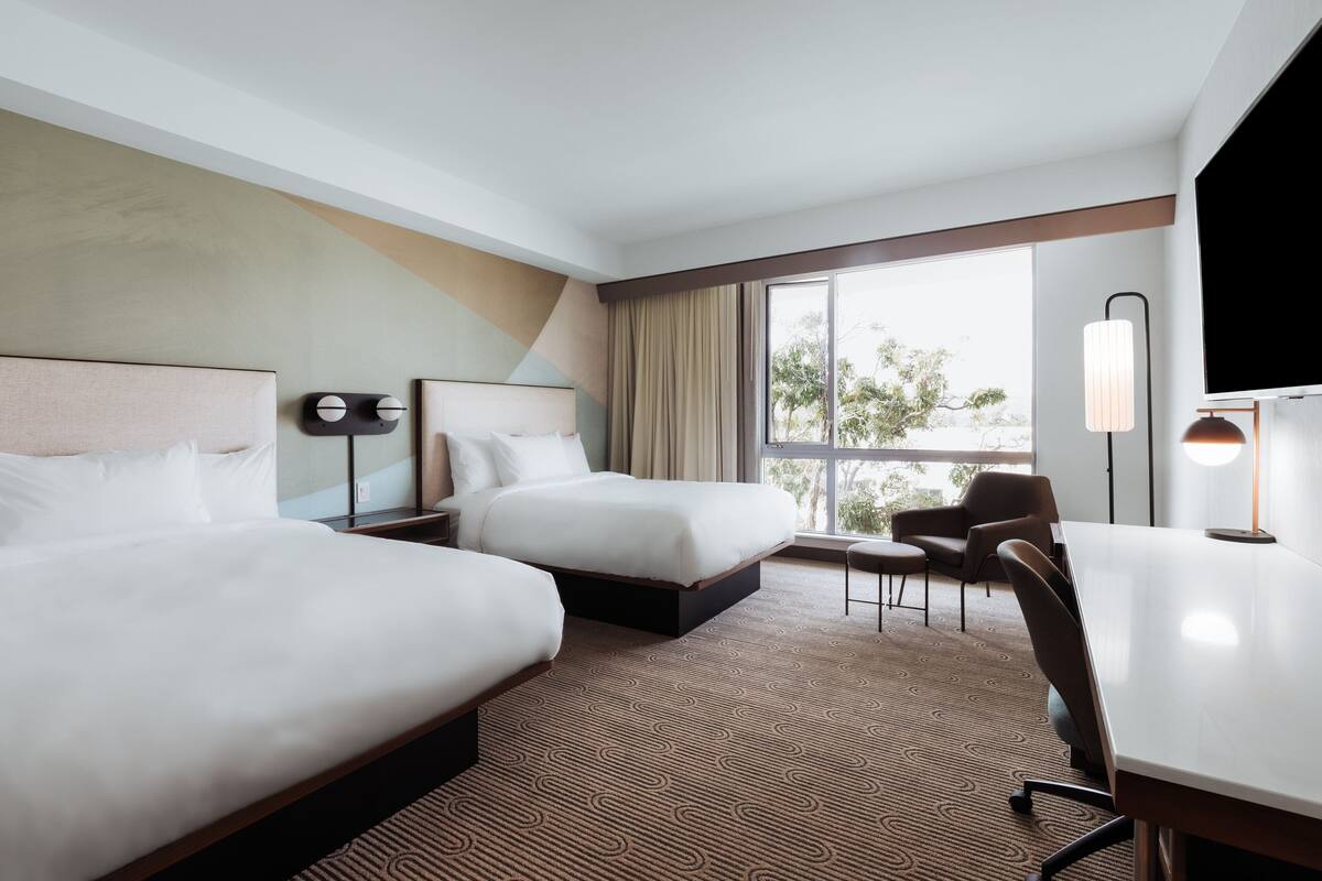 Quarto de hotel com duas camas, uma escrivaninha, uma cadeira, televisão e uma janela grande com vista.