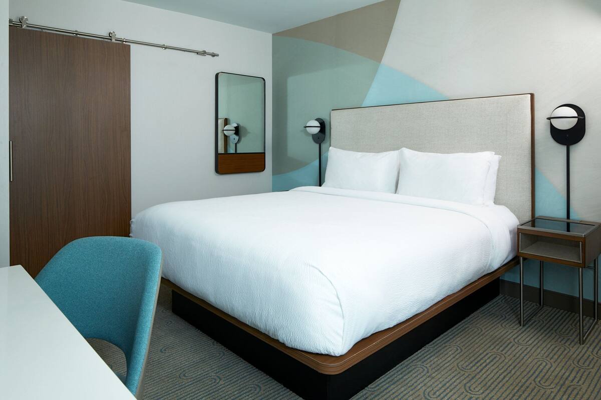 Quarto de hotel com uma cama grande, uma escrivaninha, uma cadeira, um guarda-roupa, um espelho e uma mesa de cabeceira.