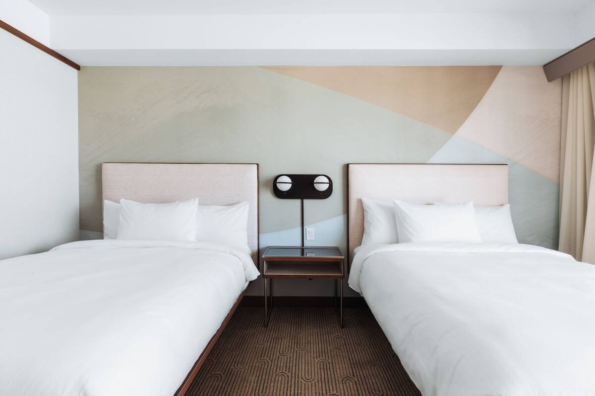 Quarto de hotel com duas camas de solteiro, uma mesa de cabeceira pequena e uma luminária fixada na parede.