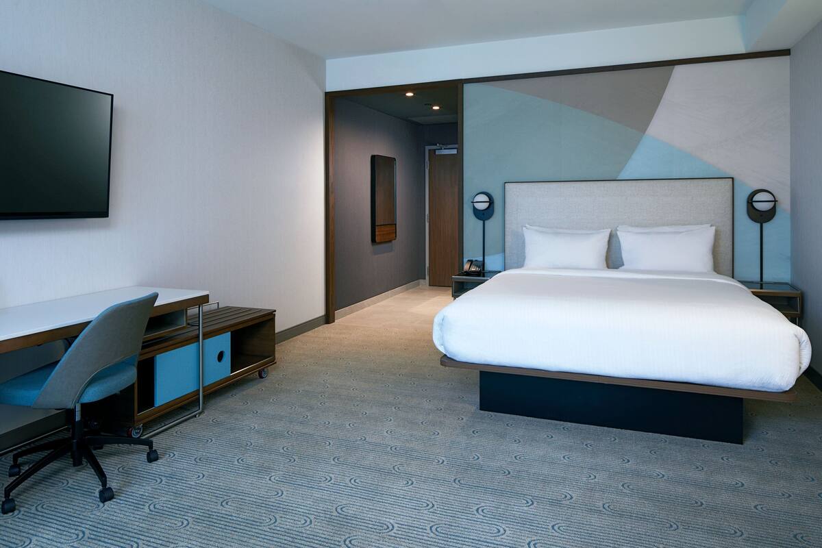Quarto de hotel moderno com uma cama grande, uma escrivaninha com cadeira e uma televisão de tela plana na parede.