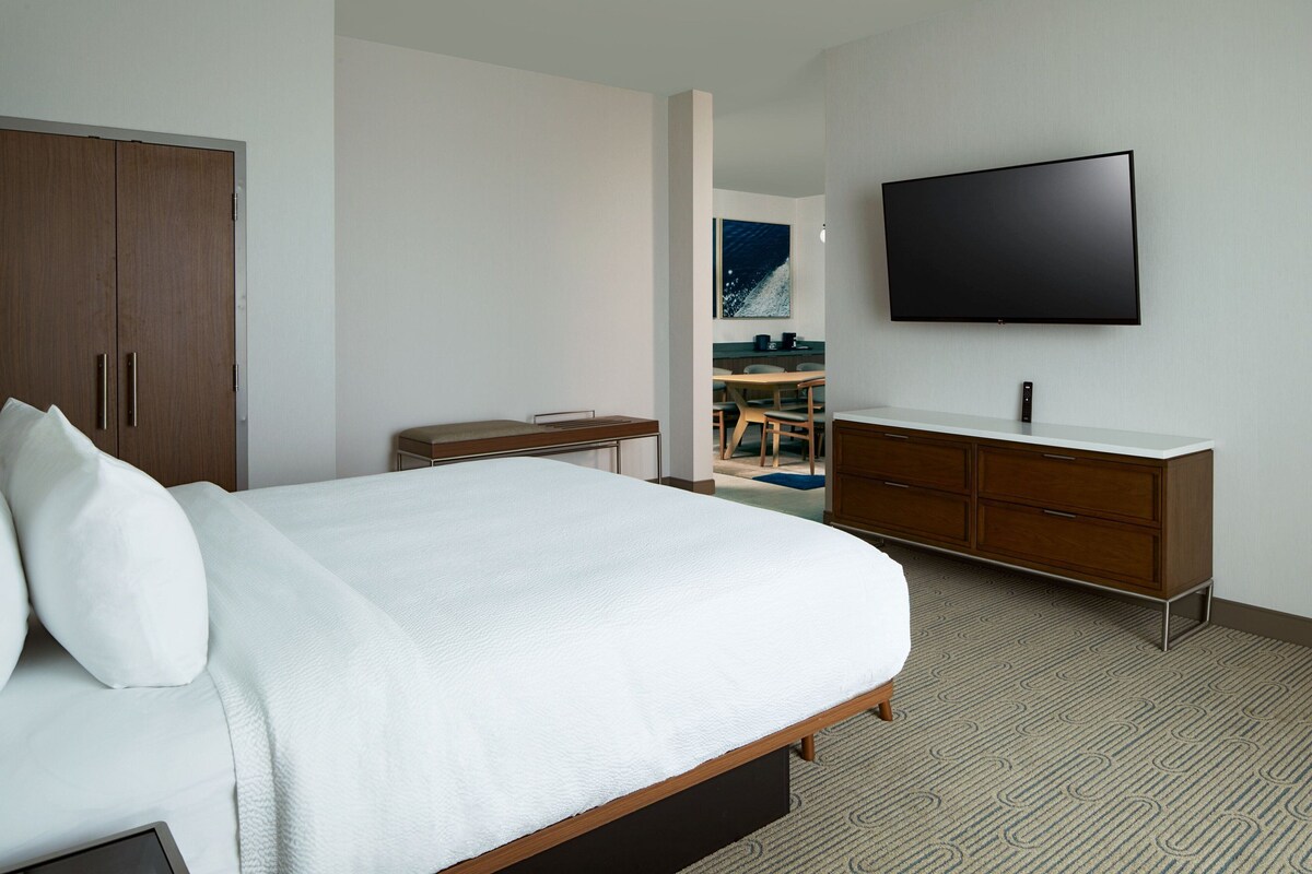 A hotel room with a large bed, a wooden wardrobe, a flat-screen TV, and a bathroom with a sink and mirror.