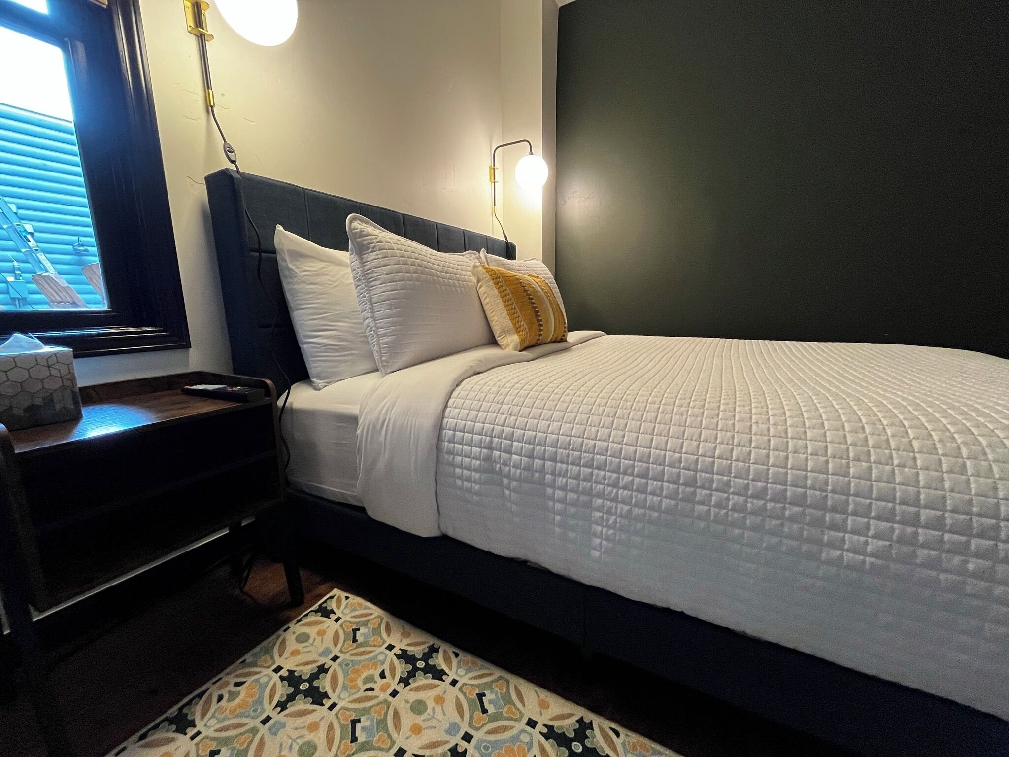 A neatly made bed with white bedding, a dark headboard, and a bedside table with a tissue box.