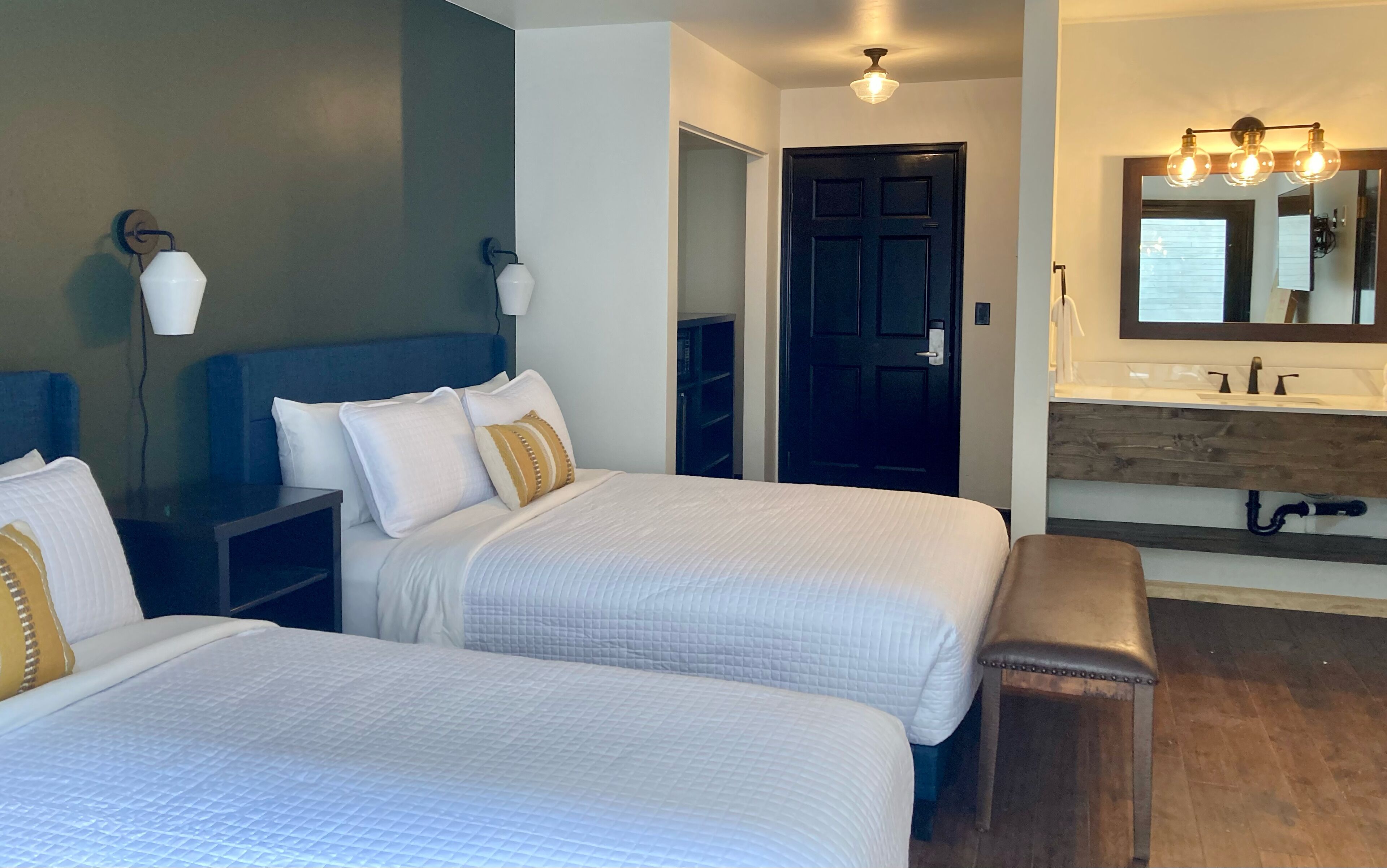A hotel room with two beds, a nightstand, a bench, a wardrobe, a door, and a bathroom with a mirror and sink.